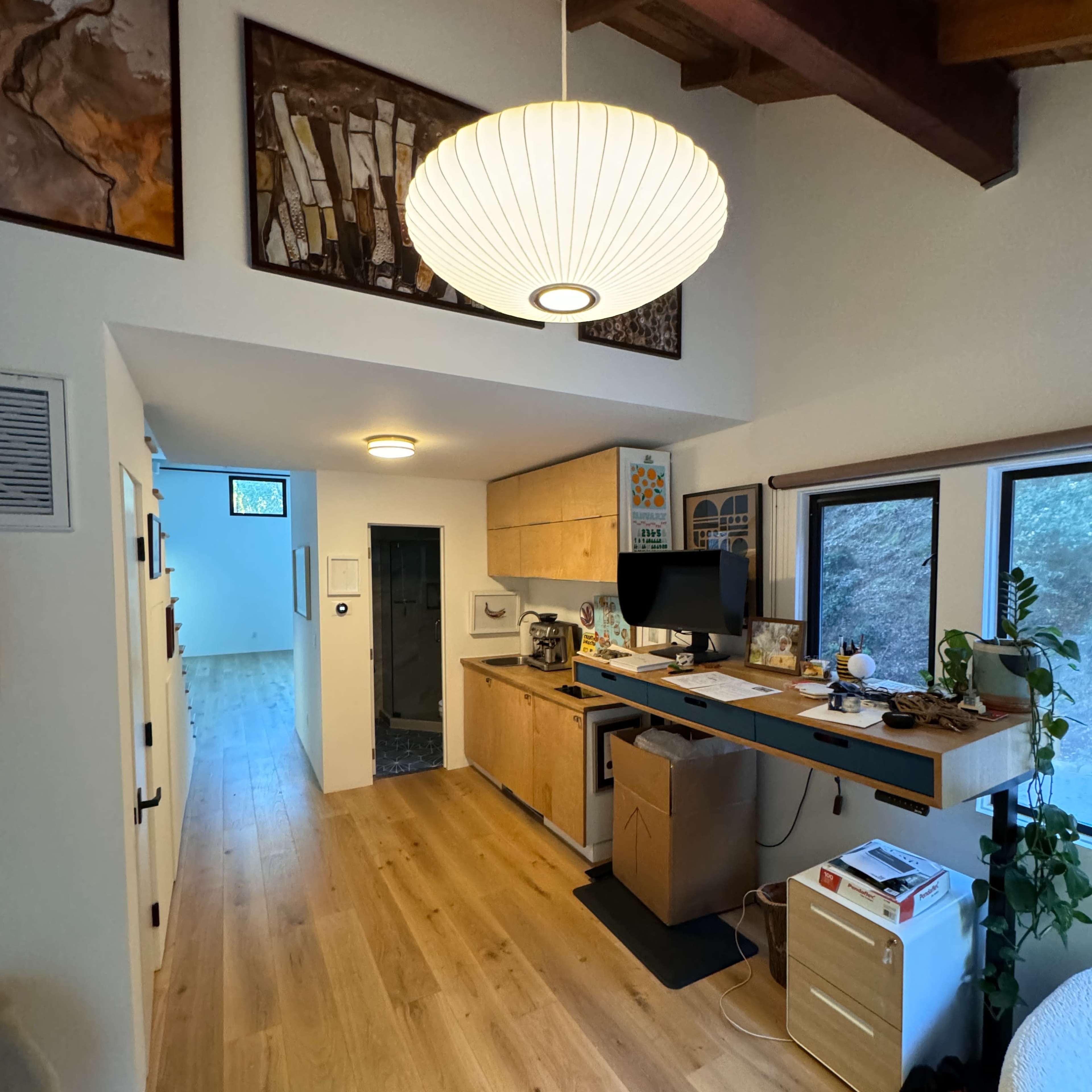 A modern home office area featuring a wooden desk, a computer setup, and a large pendant light, with a hallway and windows visible in the background.
