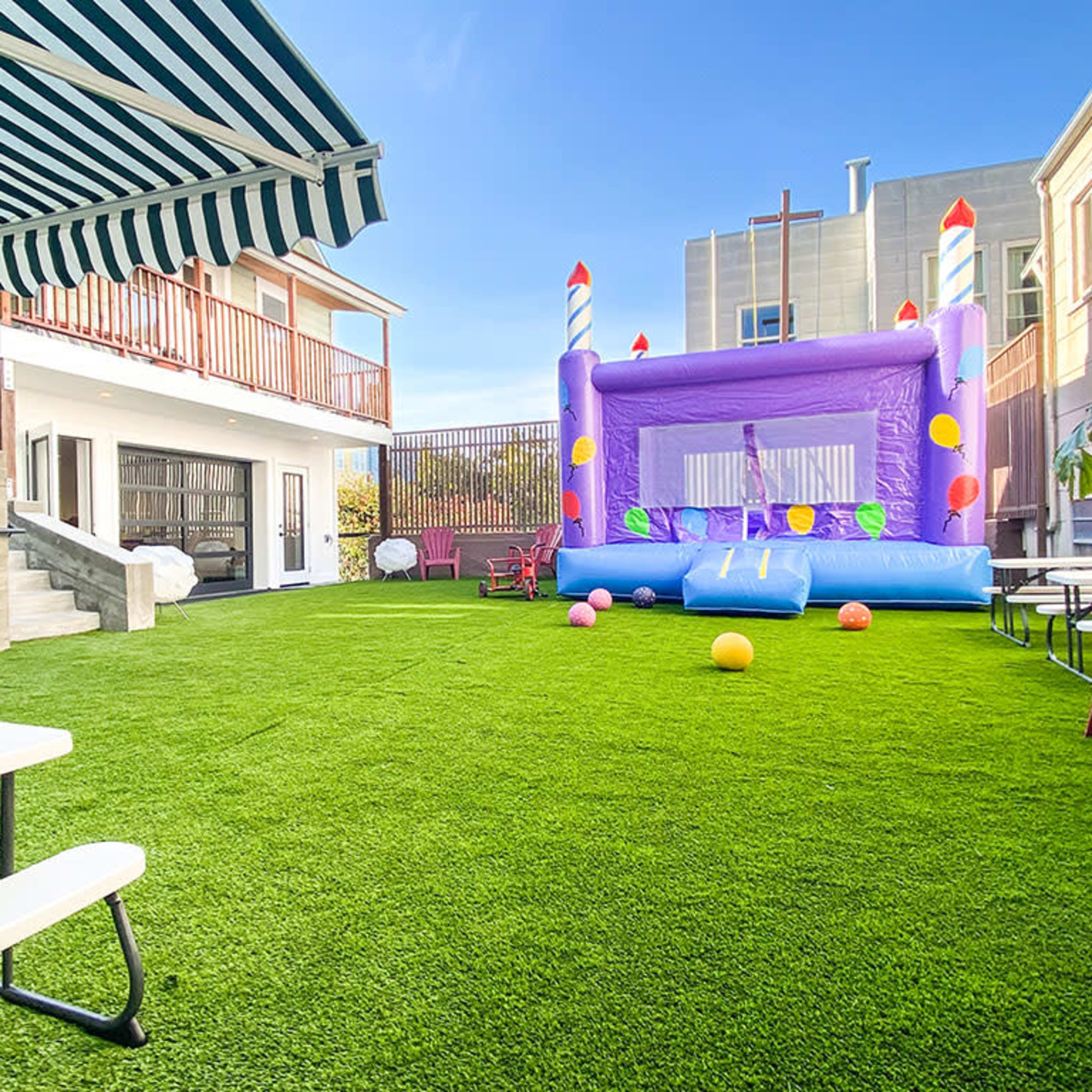 A colorful bounce house is set up on a green turf lawn next to a building with a shaded seating area.