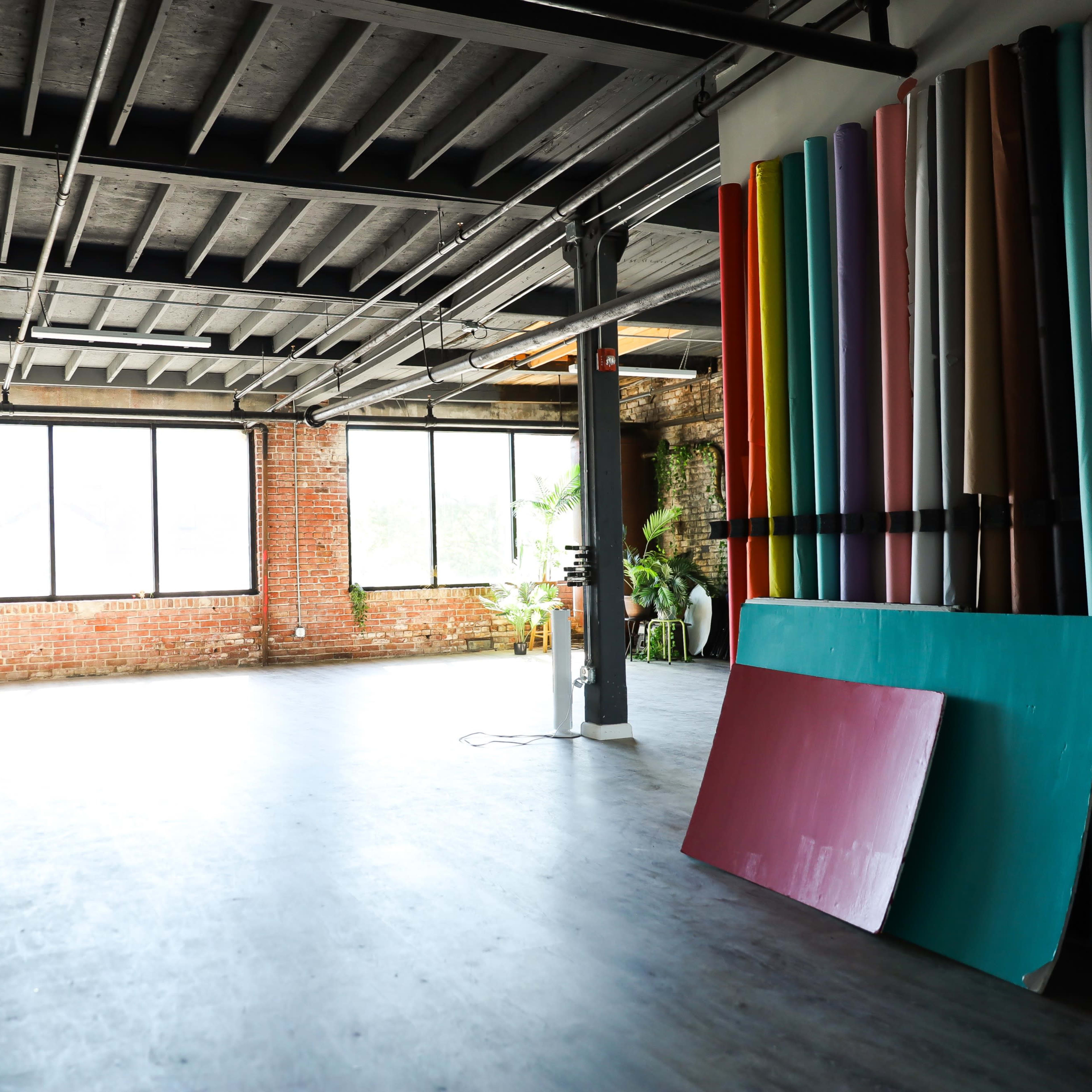 An empty studio space with large windows, exposed brick walls, and colorful panels stacked against one side.