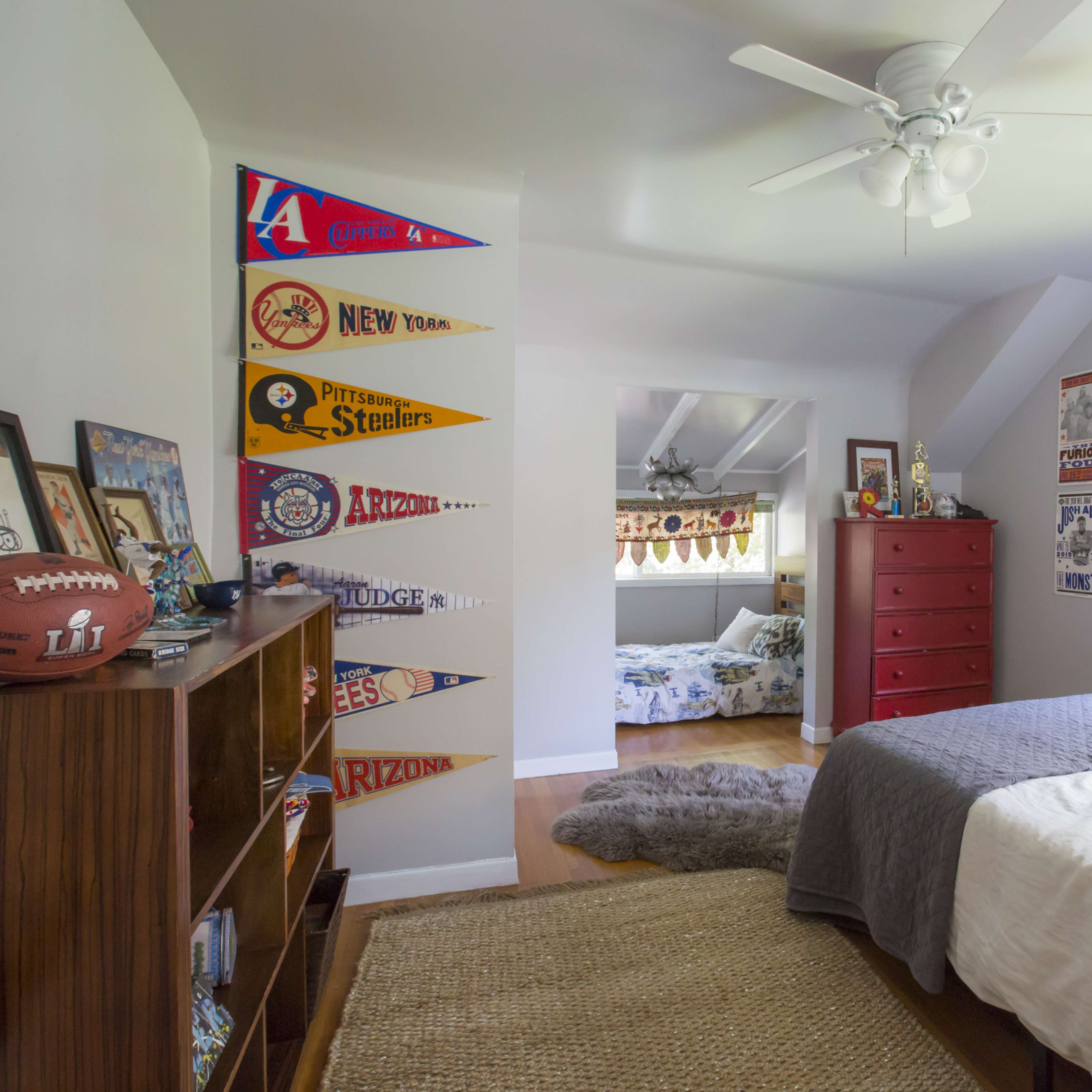 A well-lit bedroom features a bed with gray linens, sports memorabilia displayed on the walls, and a view of another room in the background.