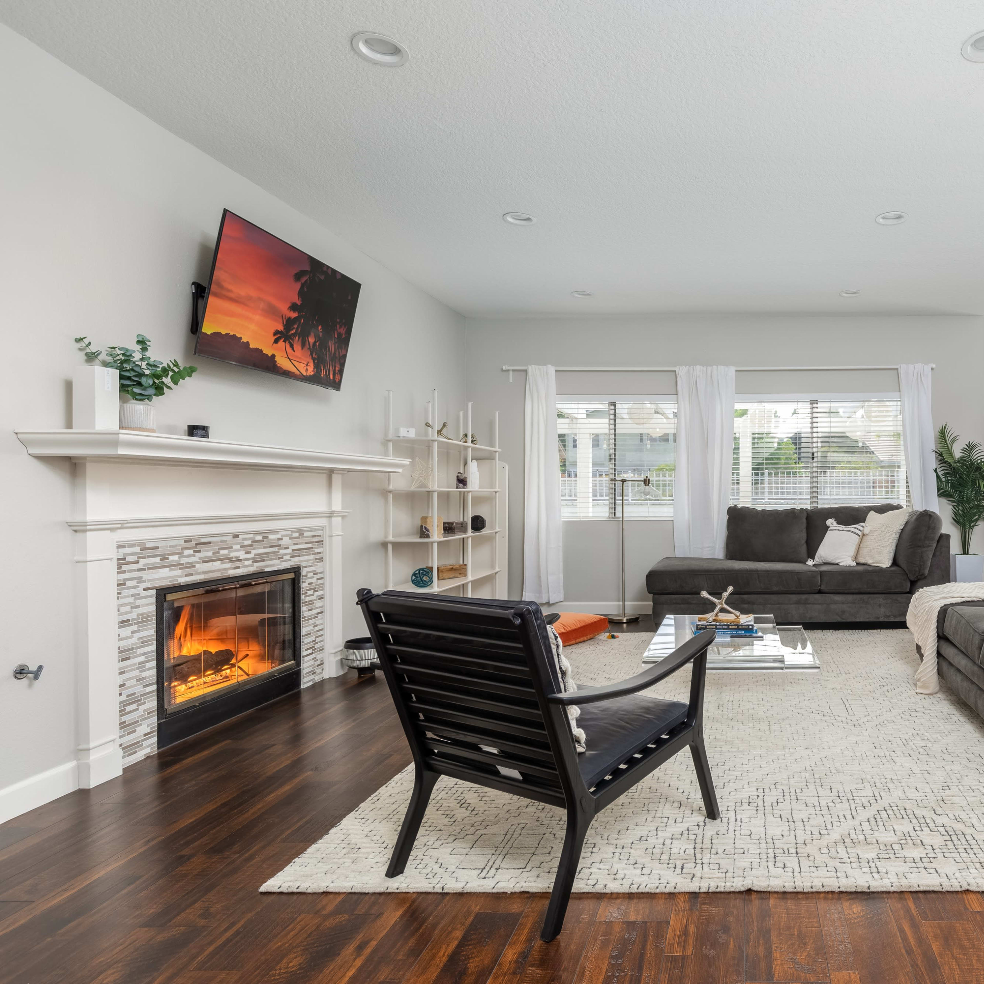 The living room features a gray sofa and loveseat, a black armchair, a coffee table, a decorative rug, and a fireplace beneath a wall-mounted television.