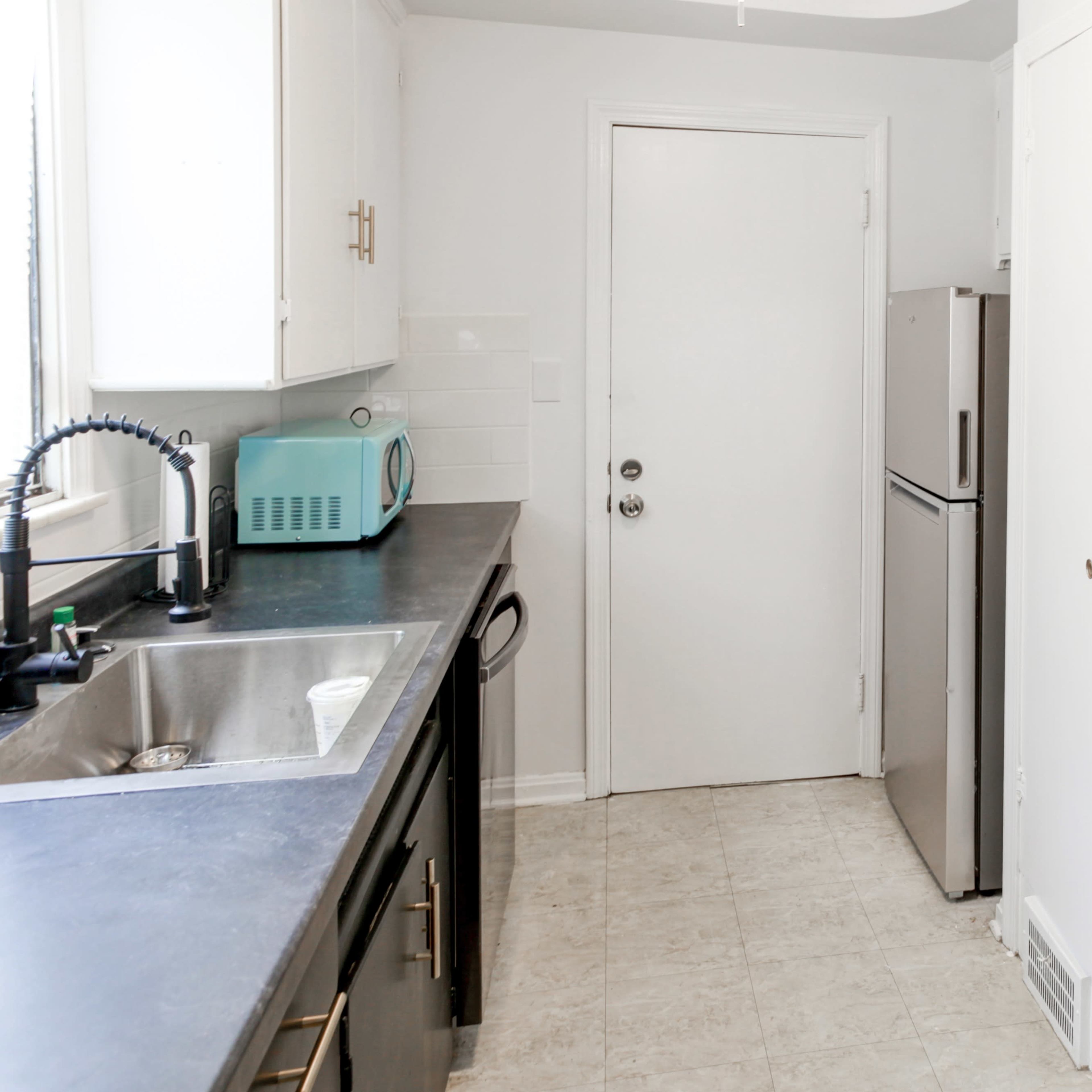 A compact kitchen featuring a sink, a stove, a refrigerator, and a door, with white cabinets and a gray countertop.