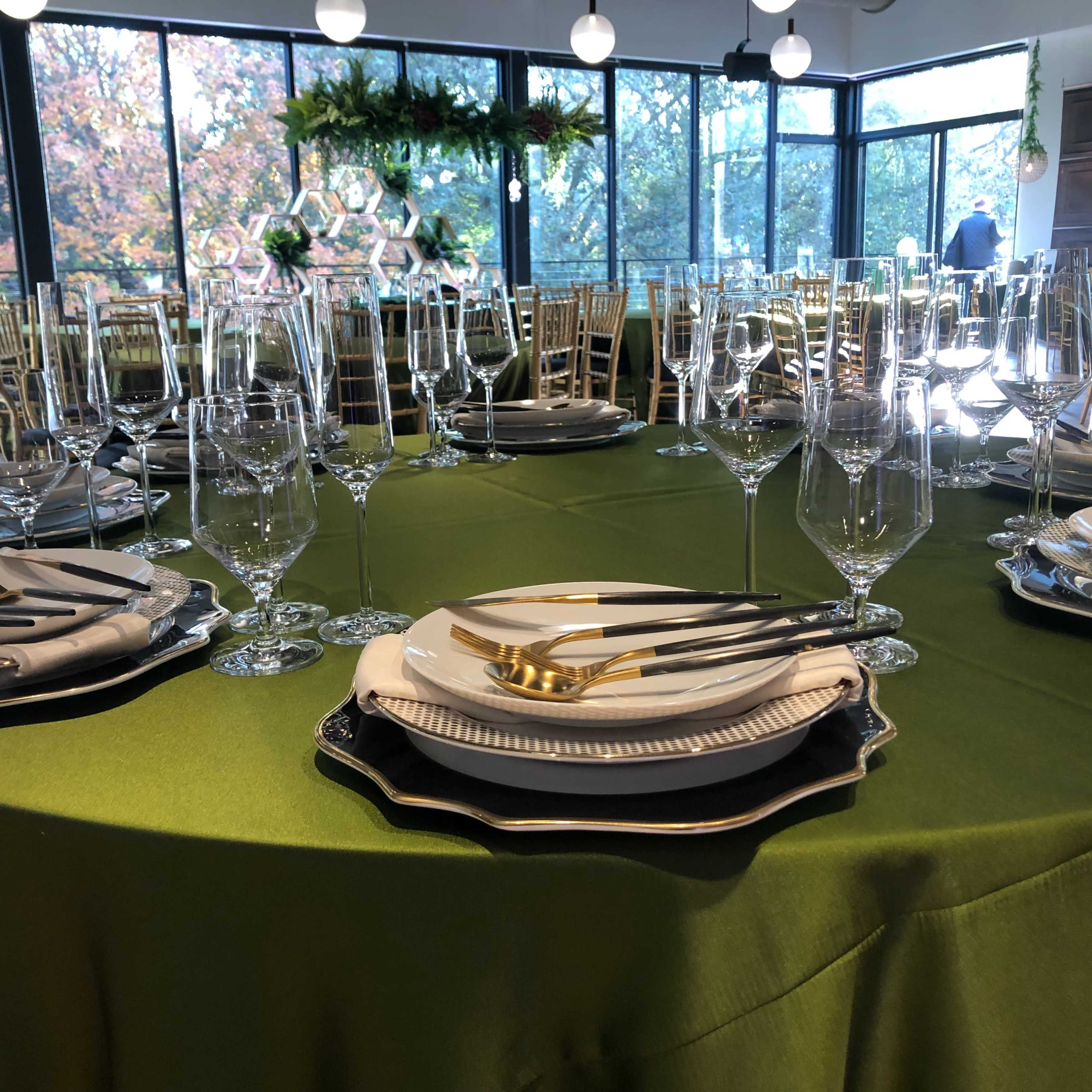 A well-lit event space with several round tables covered in green tablecloths, gold chairs, and decorative elements, along with large windows that provide an outdoor view.