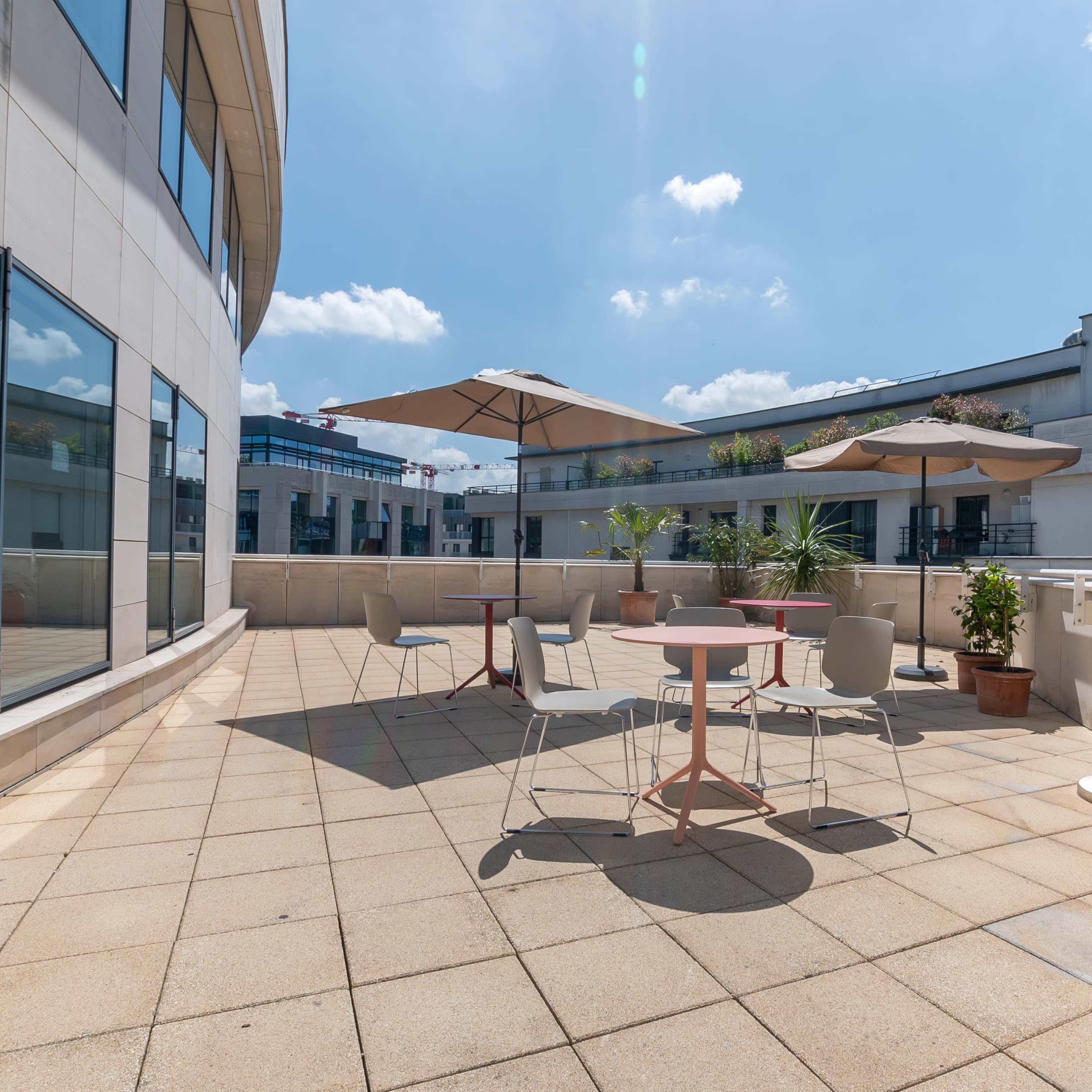 A spacious outdoor terrace with several tables and umbrellas, surrounded by windows of a modern building.