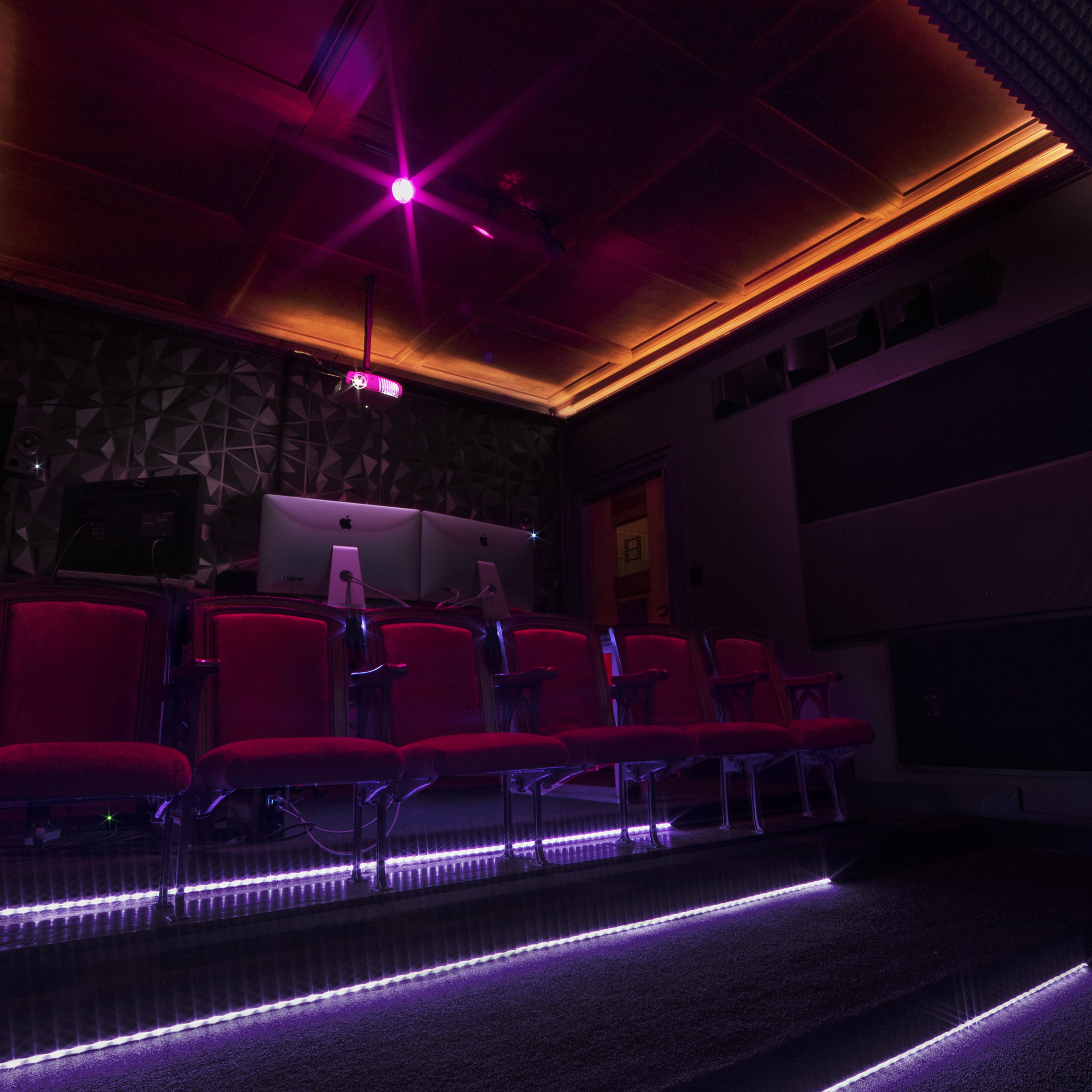 A dimly lit theater room features several red seats arranged in front of a control area, with purple lighting illuminating the space.