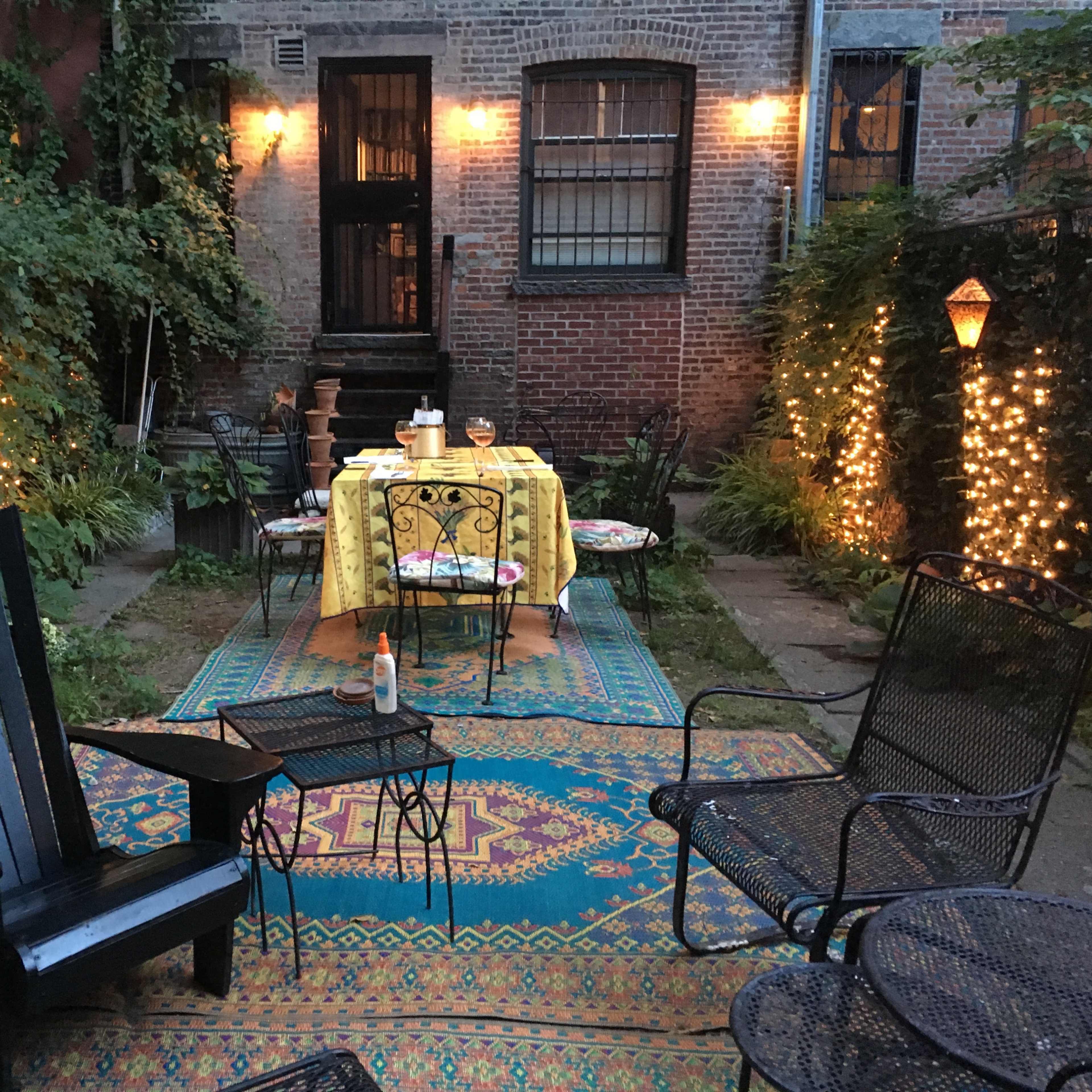 A cozy garden area with a table set for dinner, surrounded by lights and greenery, and featuring various seating options.