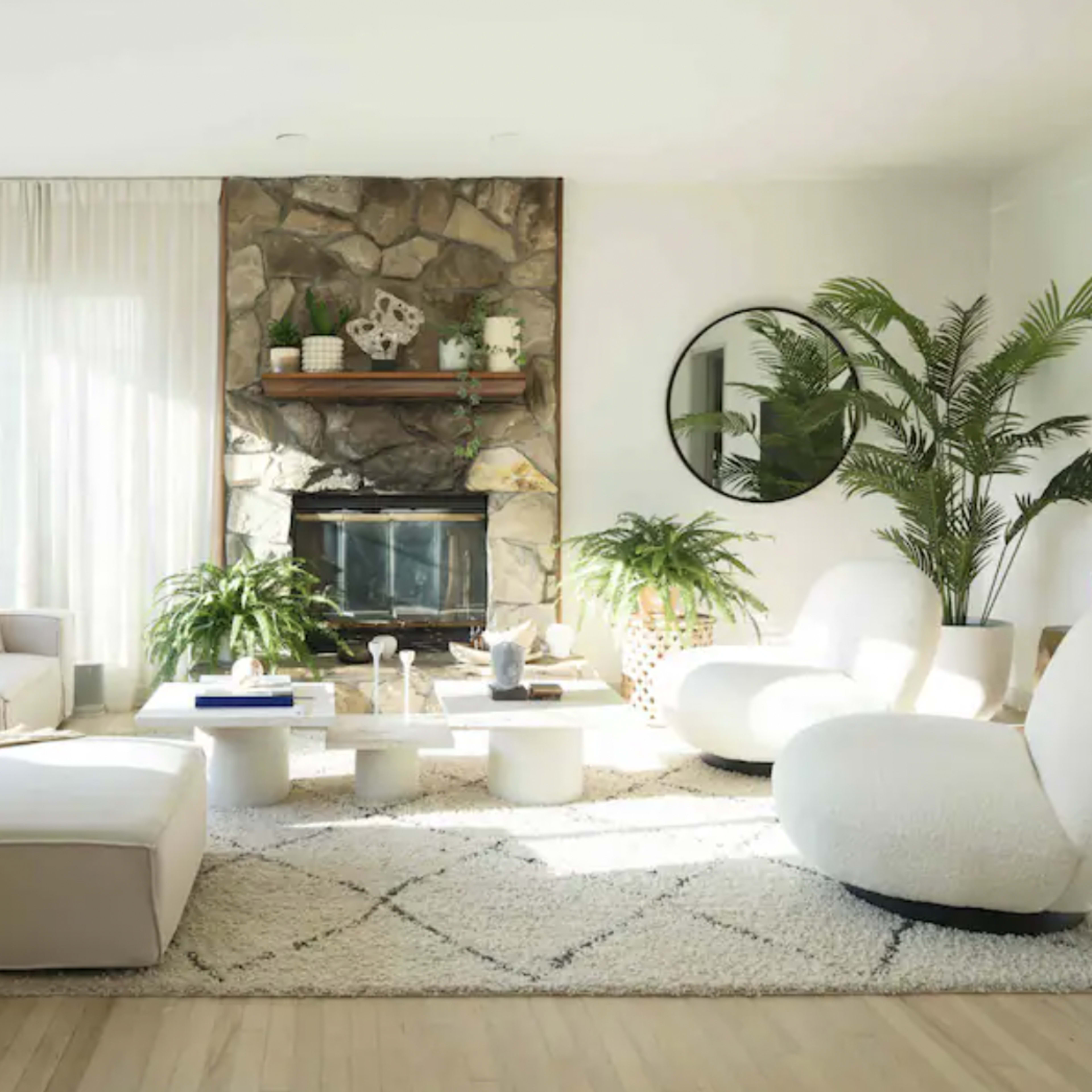 The living room features a large sectional sofa, a stone fireplace with a wooden shelf above, and potted plants scattered throughout the space, all arranged on a cream-colored area rug.