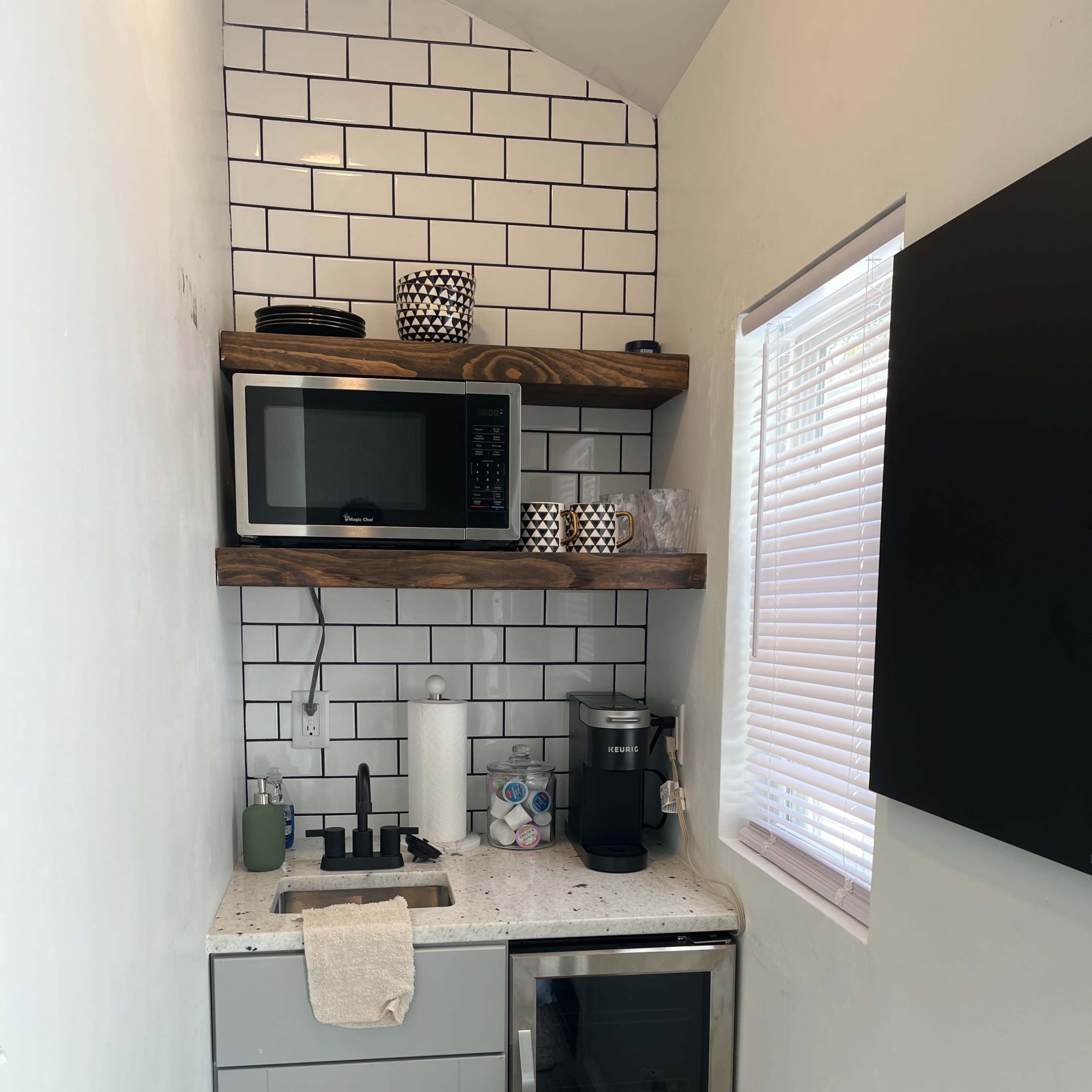 A compact kitchen area with white tiled walls, a microwave on a shelf, a coffee maker, a small refrigerator, and a sink with a modern faucet.