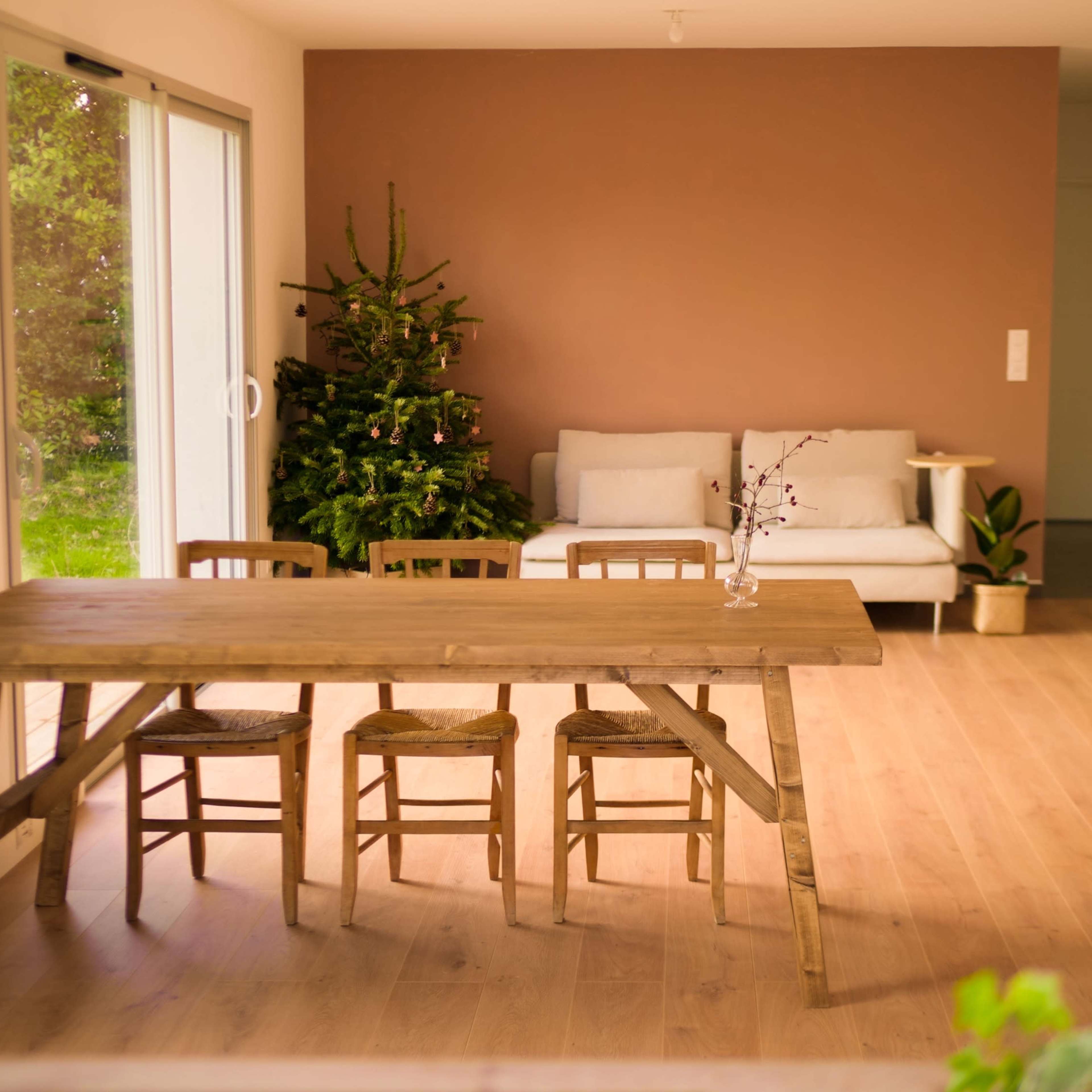 A wooden table with four chairs is set in a bright room featuring a couch, a small plant, and a Christmas tree in the background.