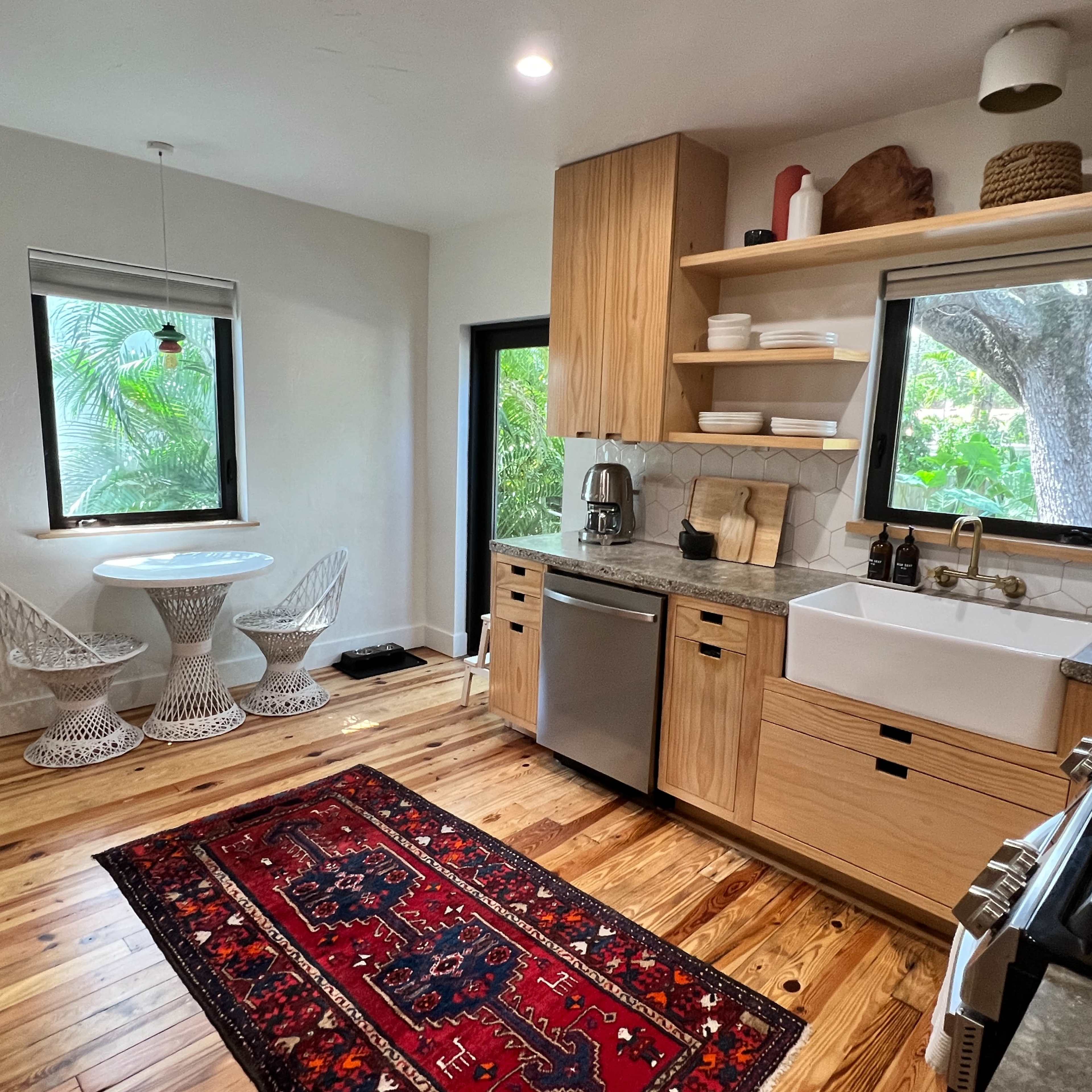 A modern kitchen features wooden cabinets, a large sink, stainless steel appliances, and a small dining area with a round table and chairs near a window overlooking greenery.