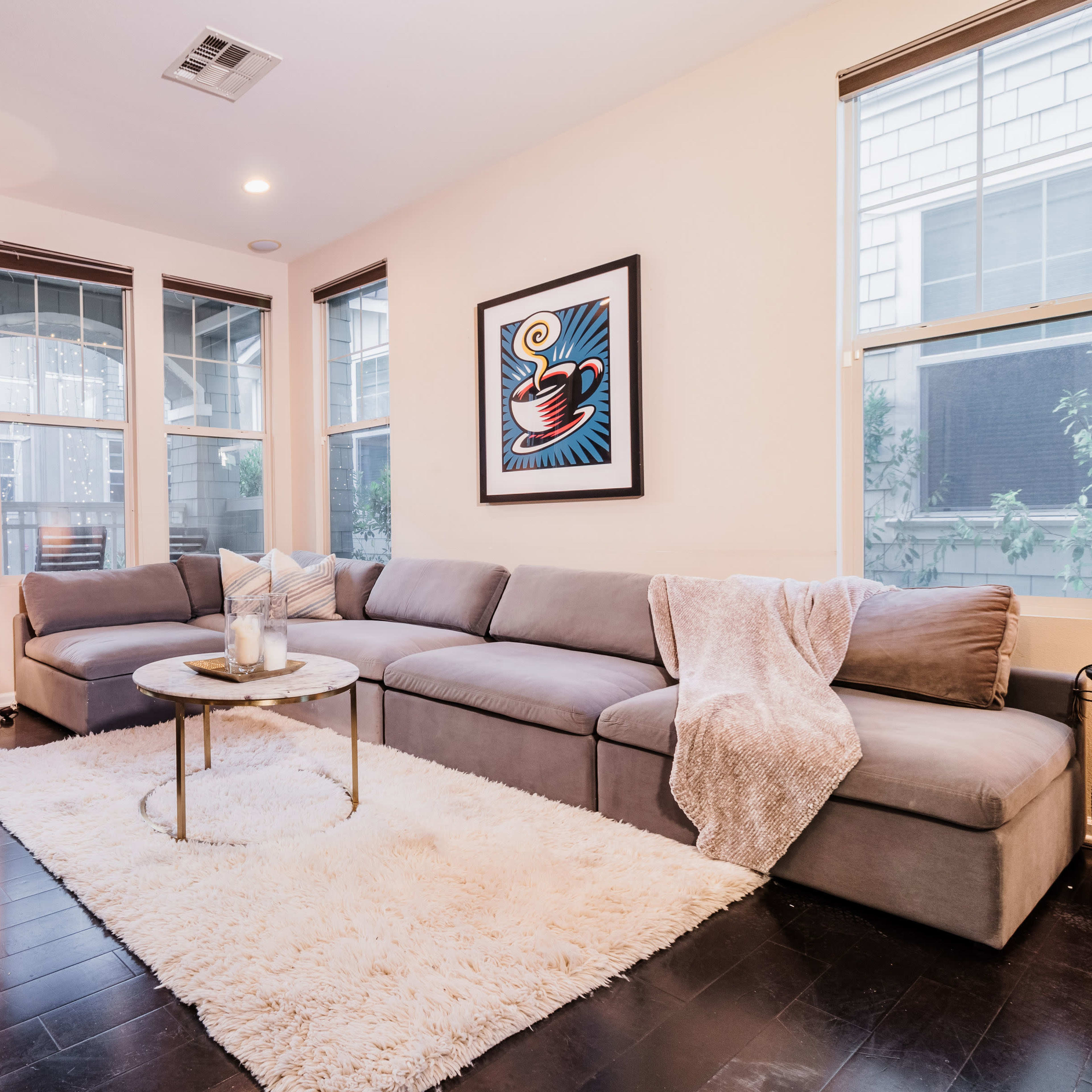 A modern living room with a gray sectional sofa, a round coffee table on a white rug, and large windows letting in natural light.