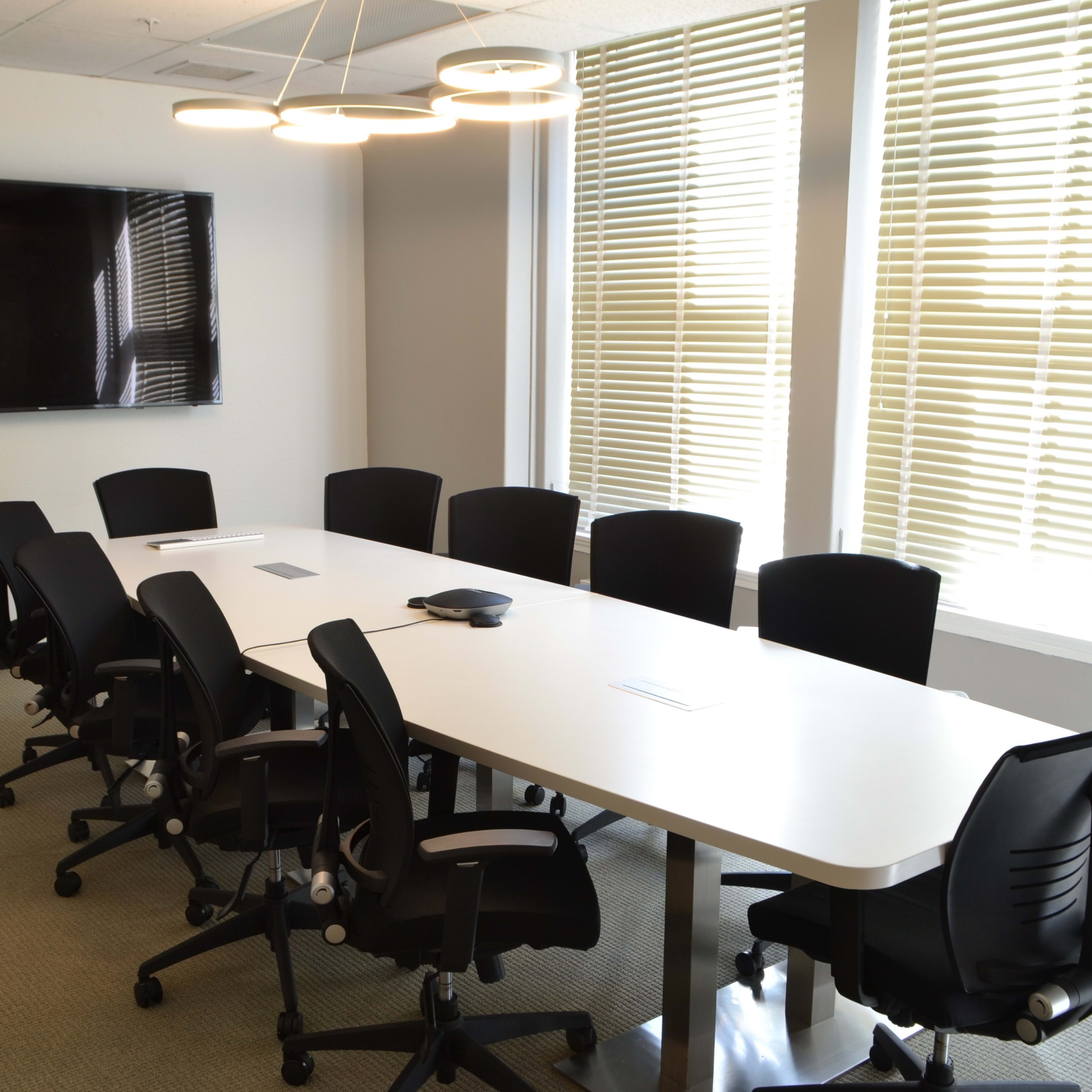A modern conference room features a long white table surrounded by black ergonomic chairs, with a large TV on the wall and natural light filtering through window blinds.