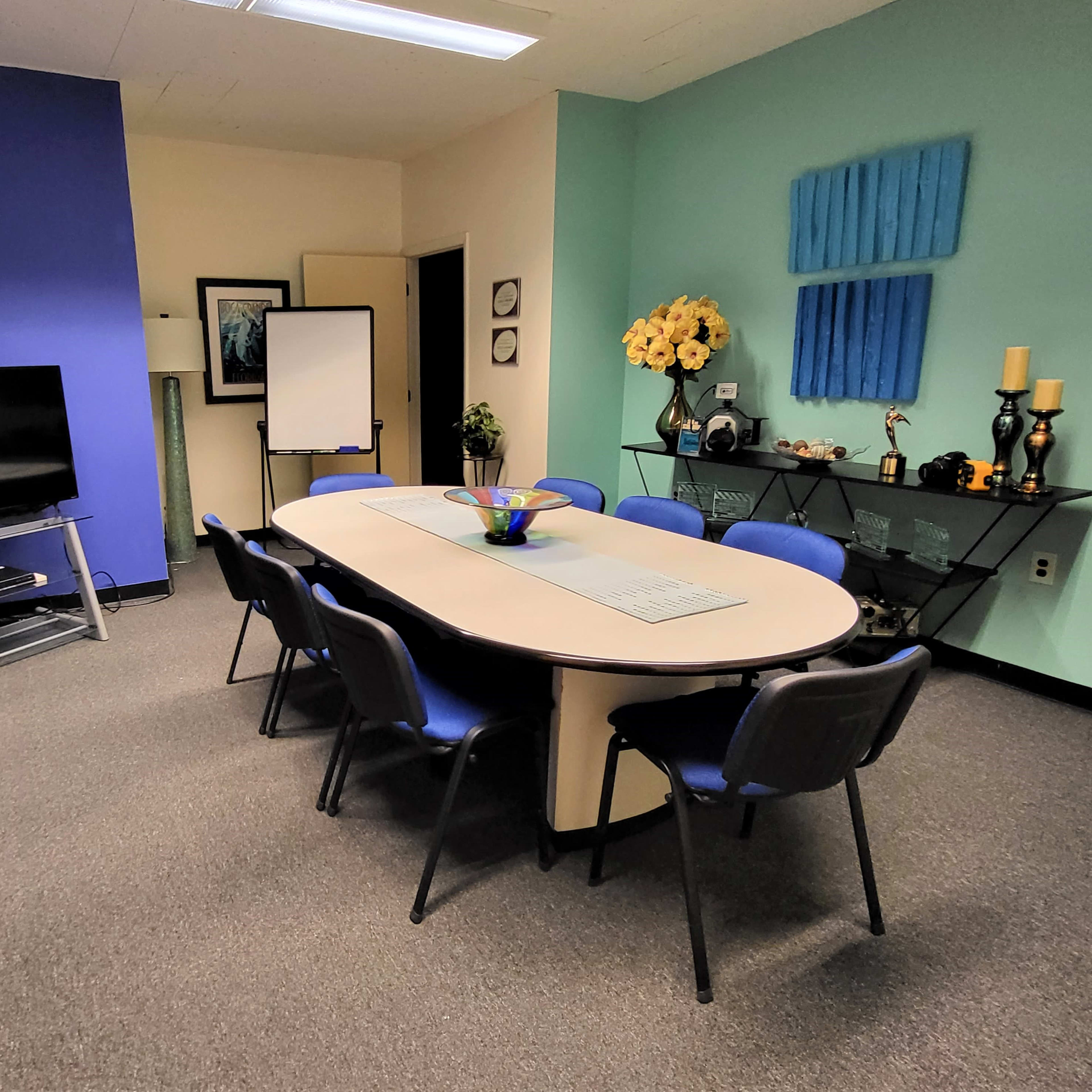 A meeting room with a large oval table surrounded by blue chairs, a television on a stand, and decorative artwork on the walls.