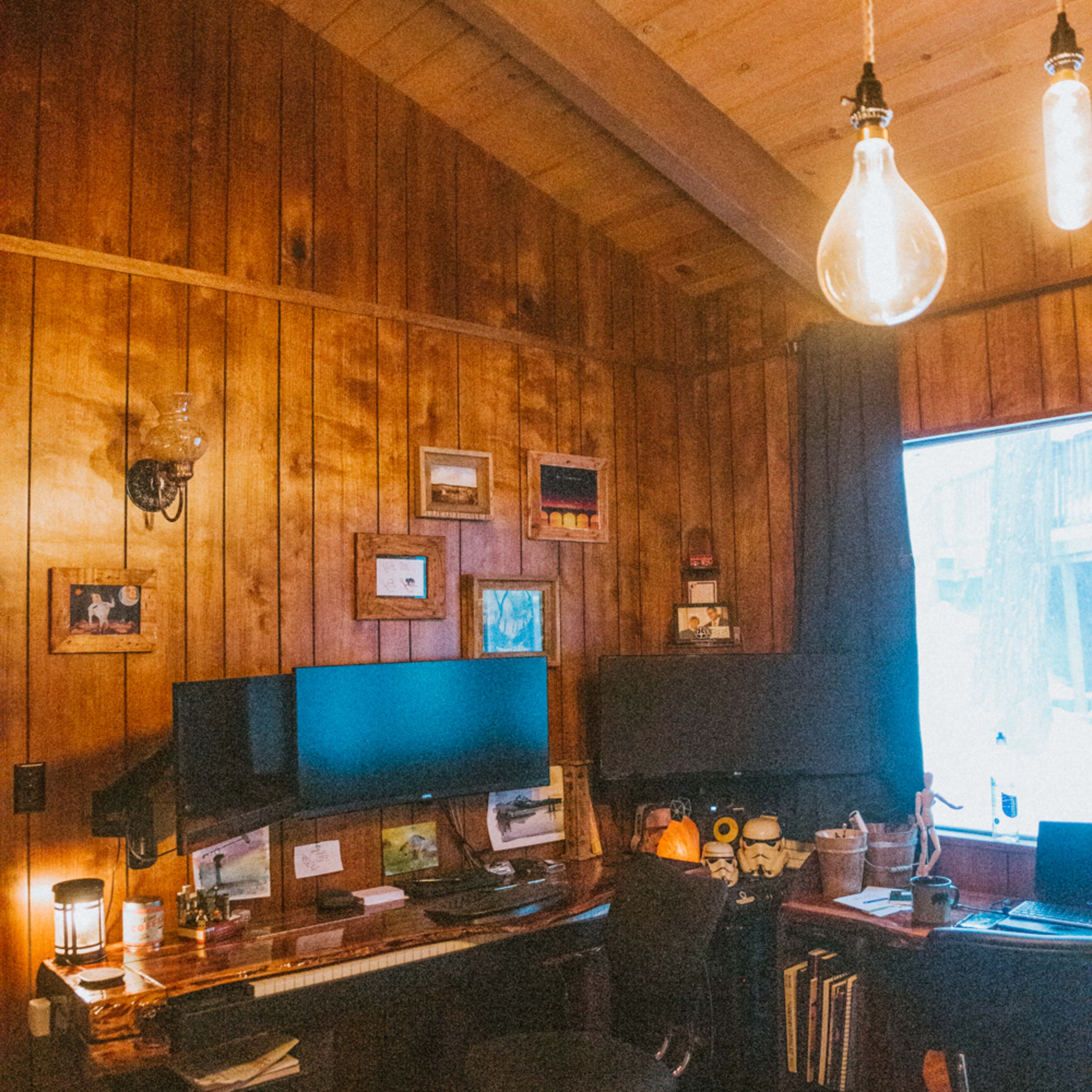 A wooden-paneled office with dual computer monitors, an ergonomic chair, and warm lighting from hanging bulbs.