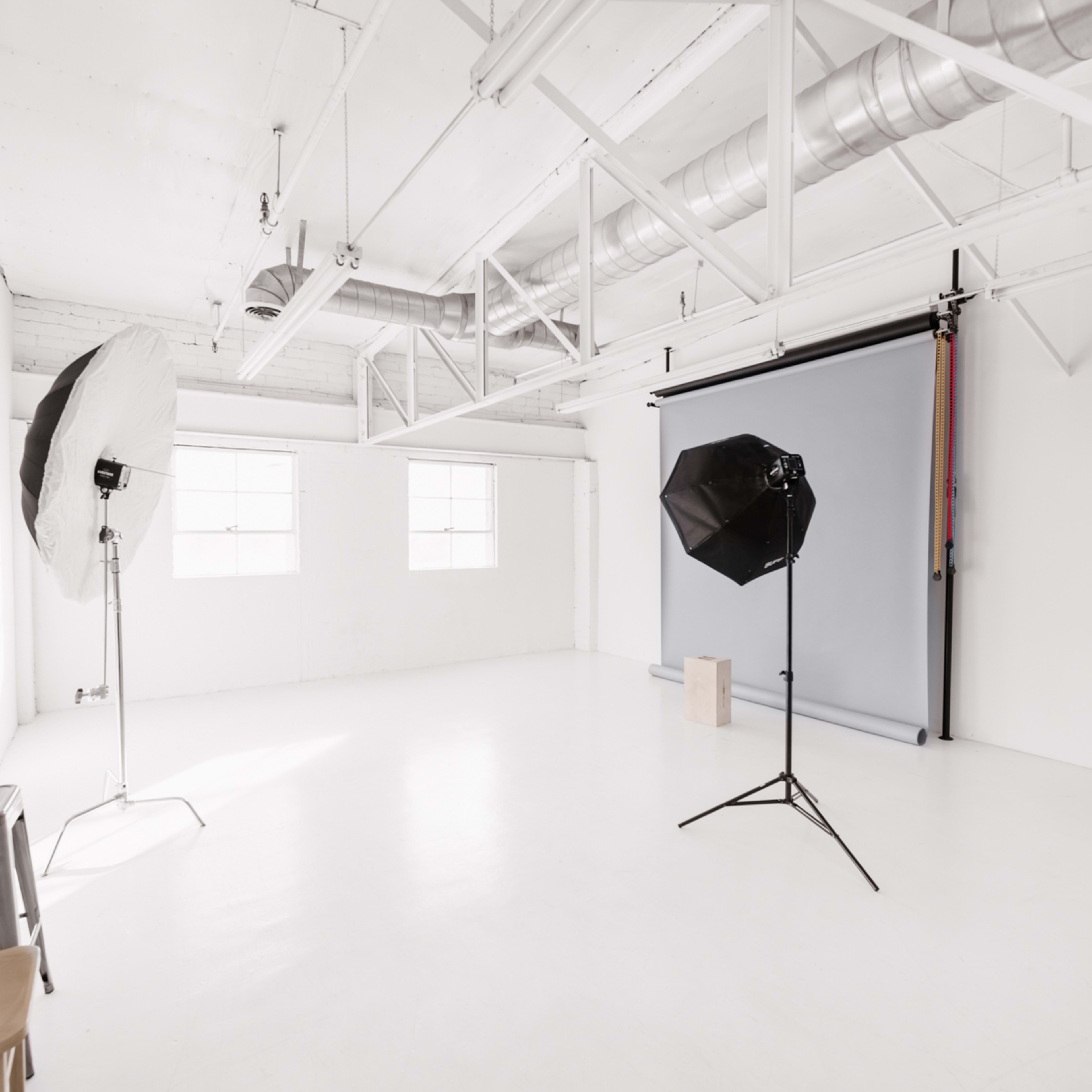 A spacious, well-lit photography studio with two light setup stands, a backdrop, and a few chairs.