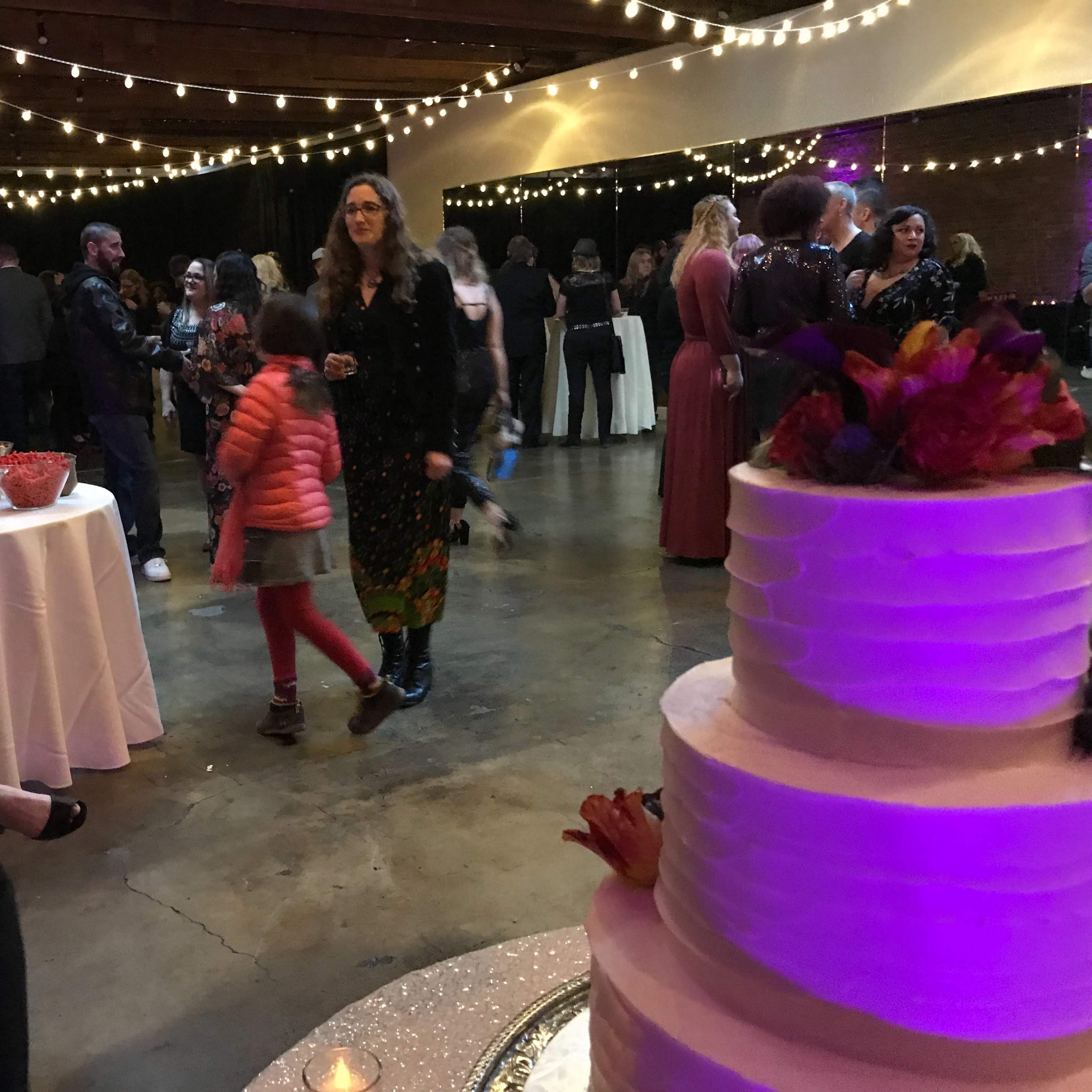 A large indoor event space is filled with guests, tables of food, and a decorated cake, illuminated by string lights overhead.