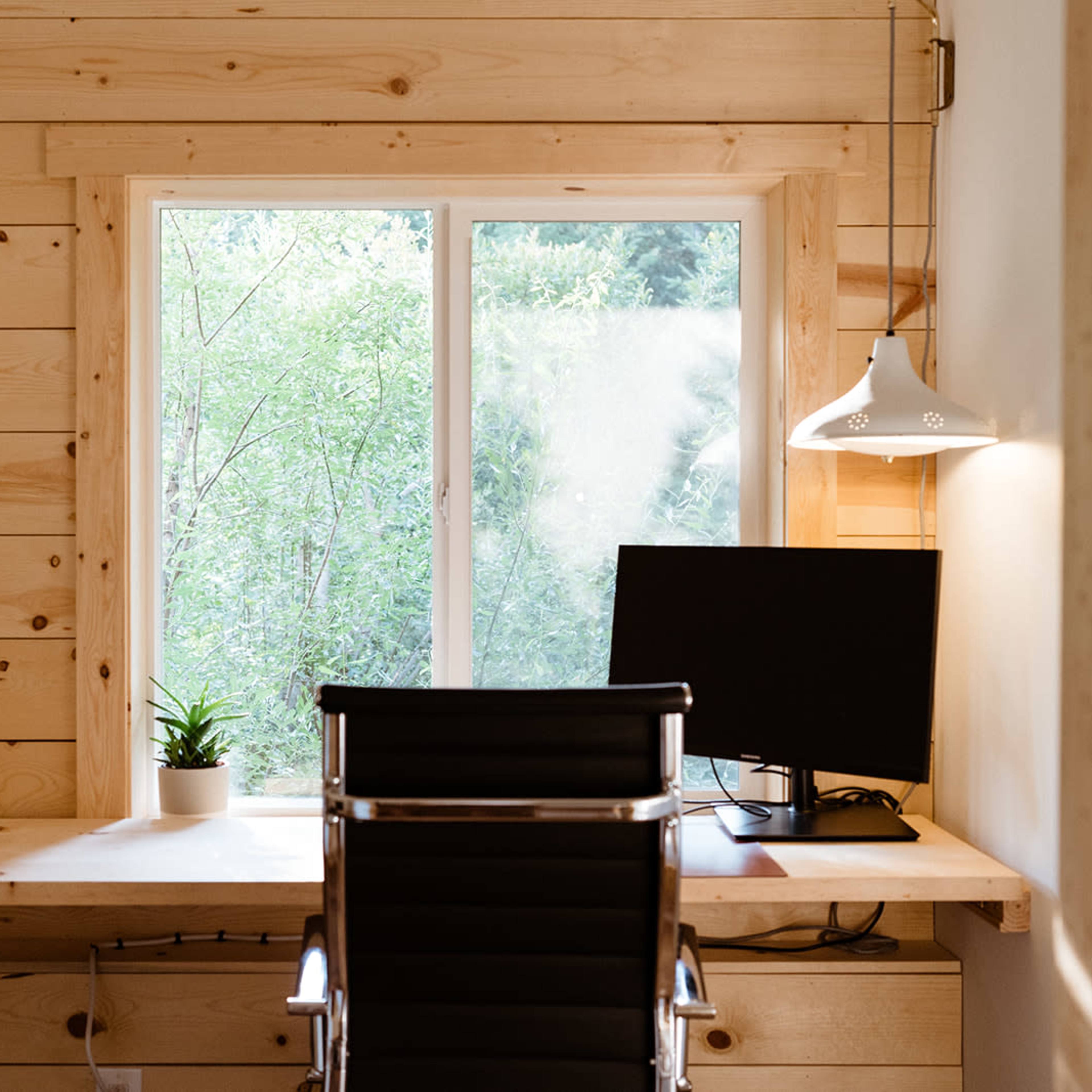 A minimalist home office features a desk with a computer and a chair, positioned beside a large window overlooking greenery.