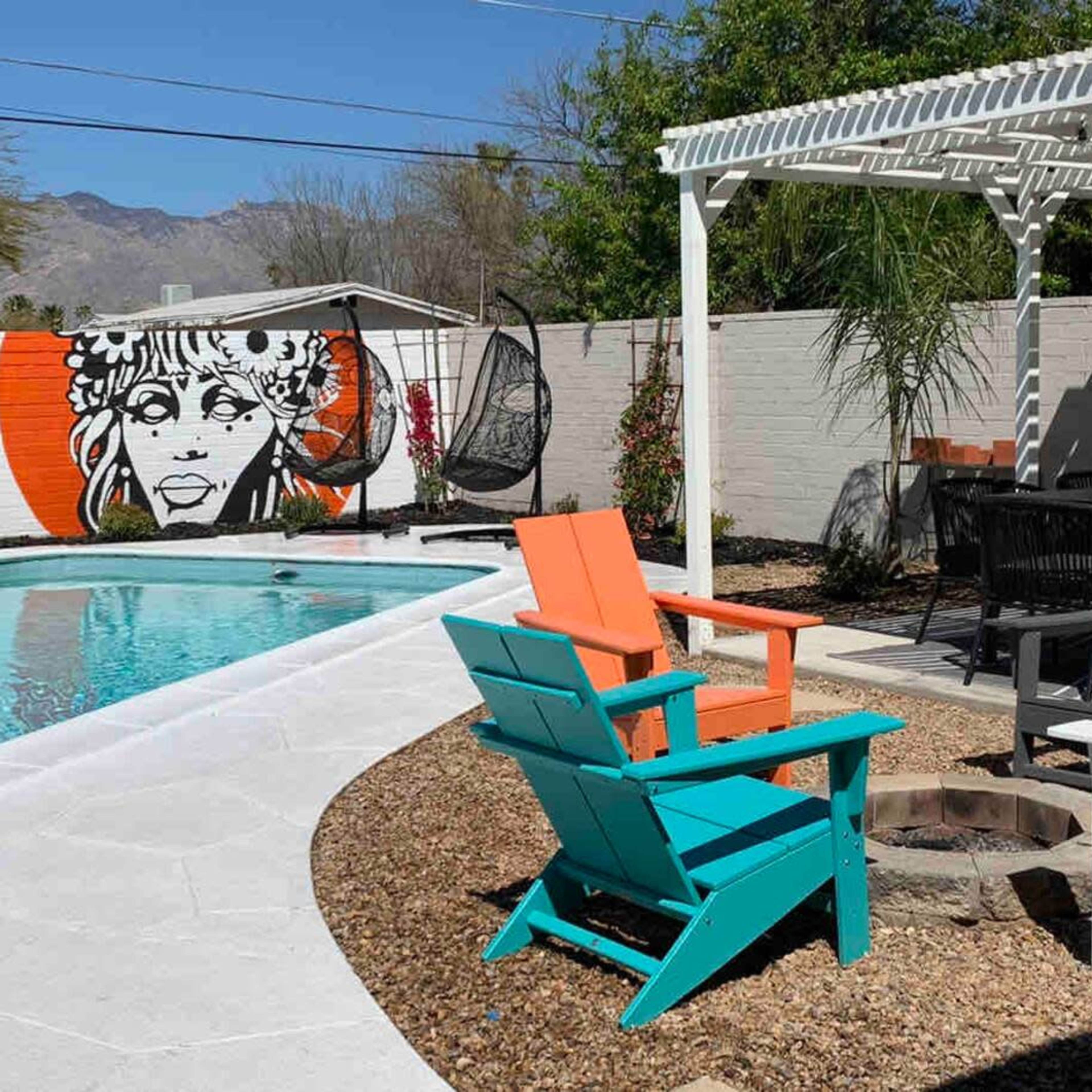 A brightly colored backyard pool area with lounge chairs, a fire pit, and a mural of a woman's face on the wall.
