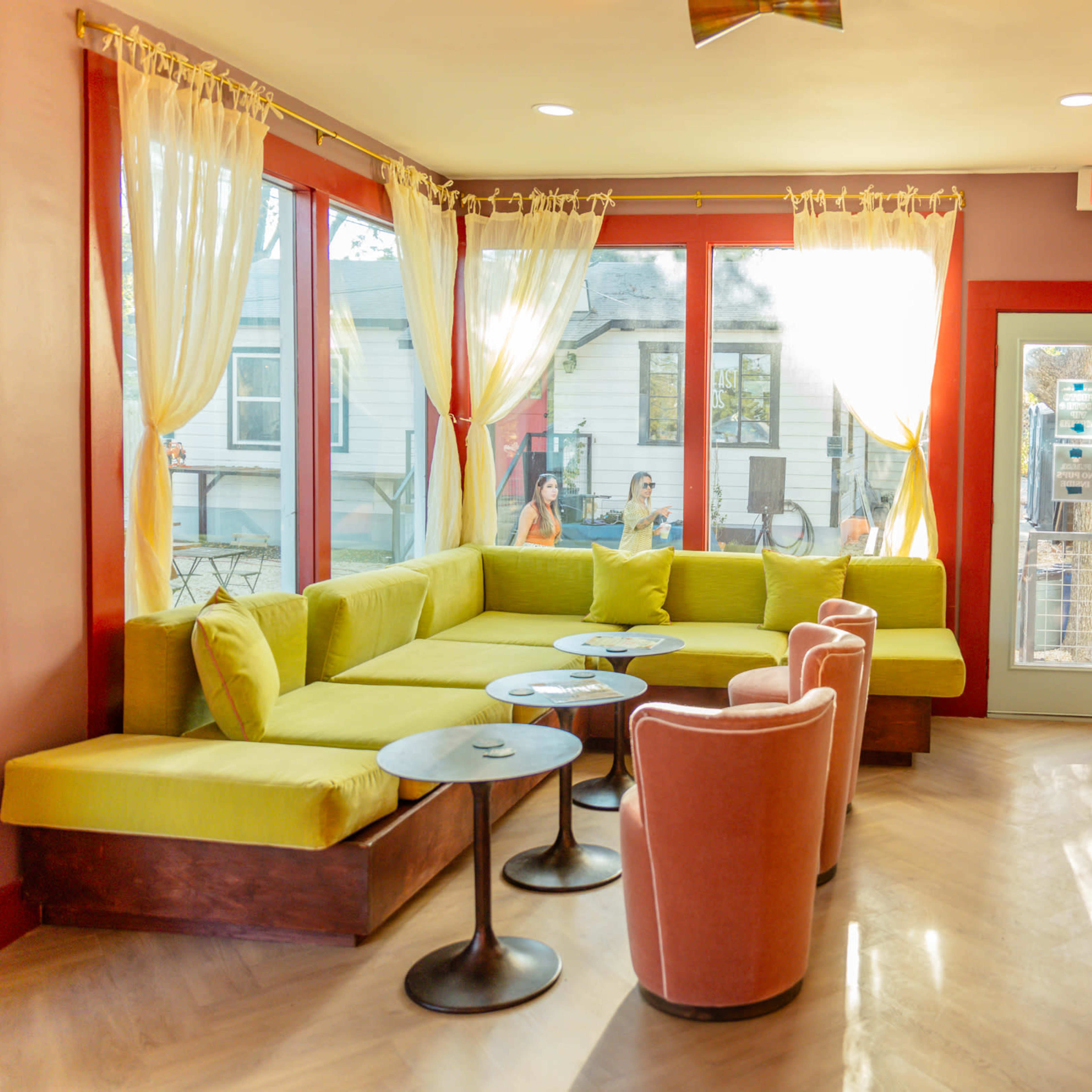 A bright, modern seating area features a large green sectional, pink chairs, and round tables, surrounded by windows with sheer curtains.