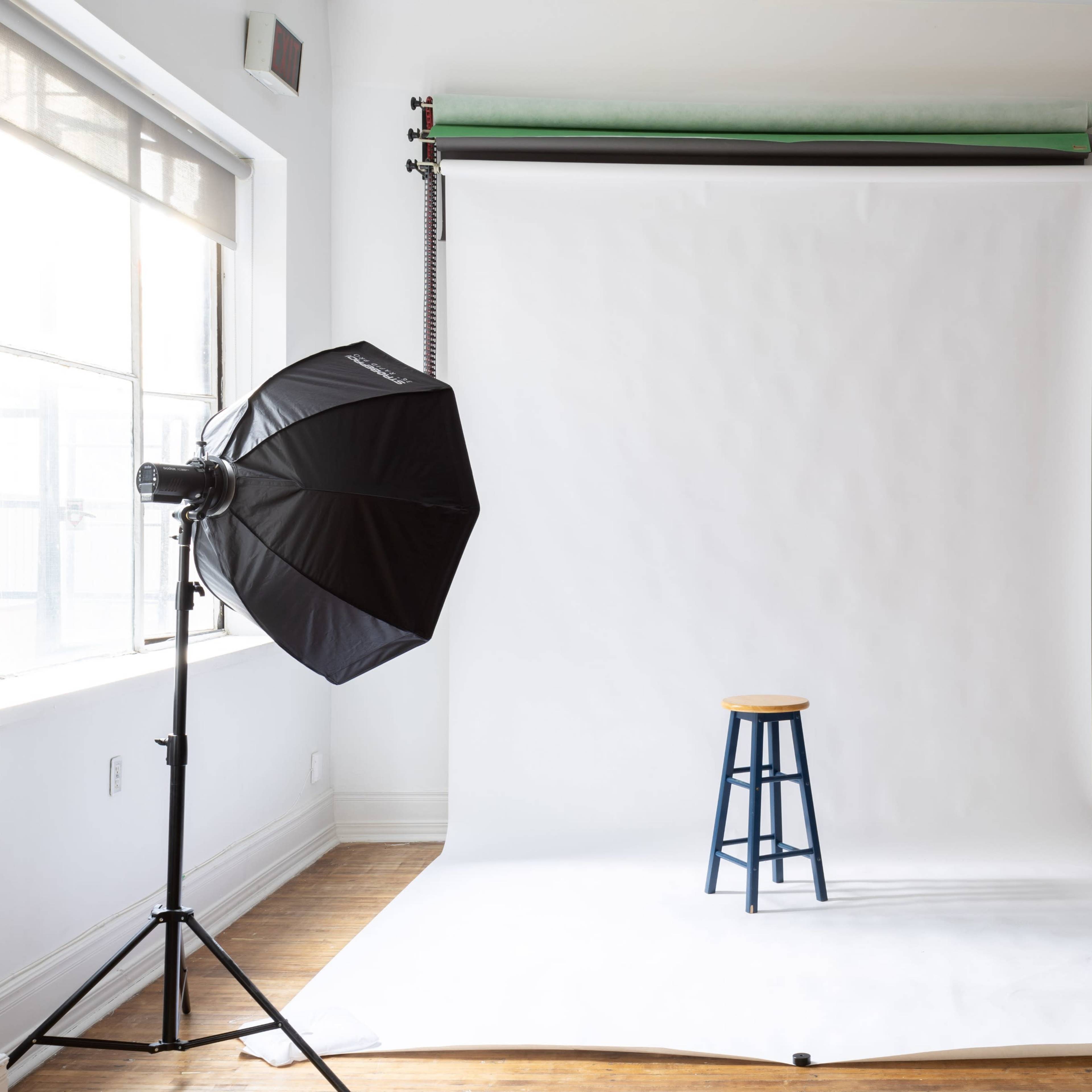 A photography studio with a white backdrop, two light sources, and a wooden stool in the center.