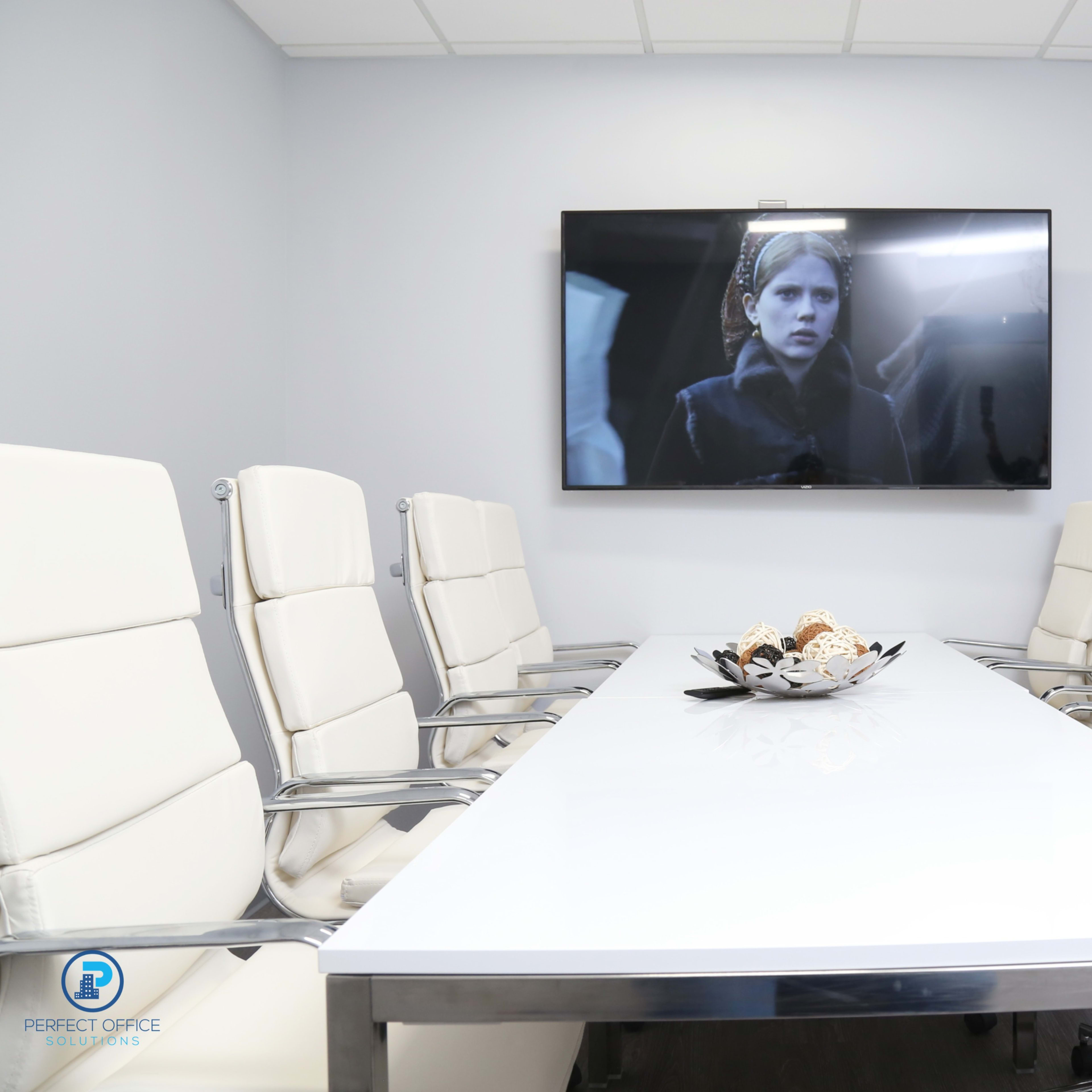 A modern conference room features a long white table surrounded by eight white chairs and a large television screen mounted on the wall.
