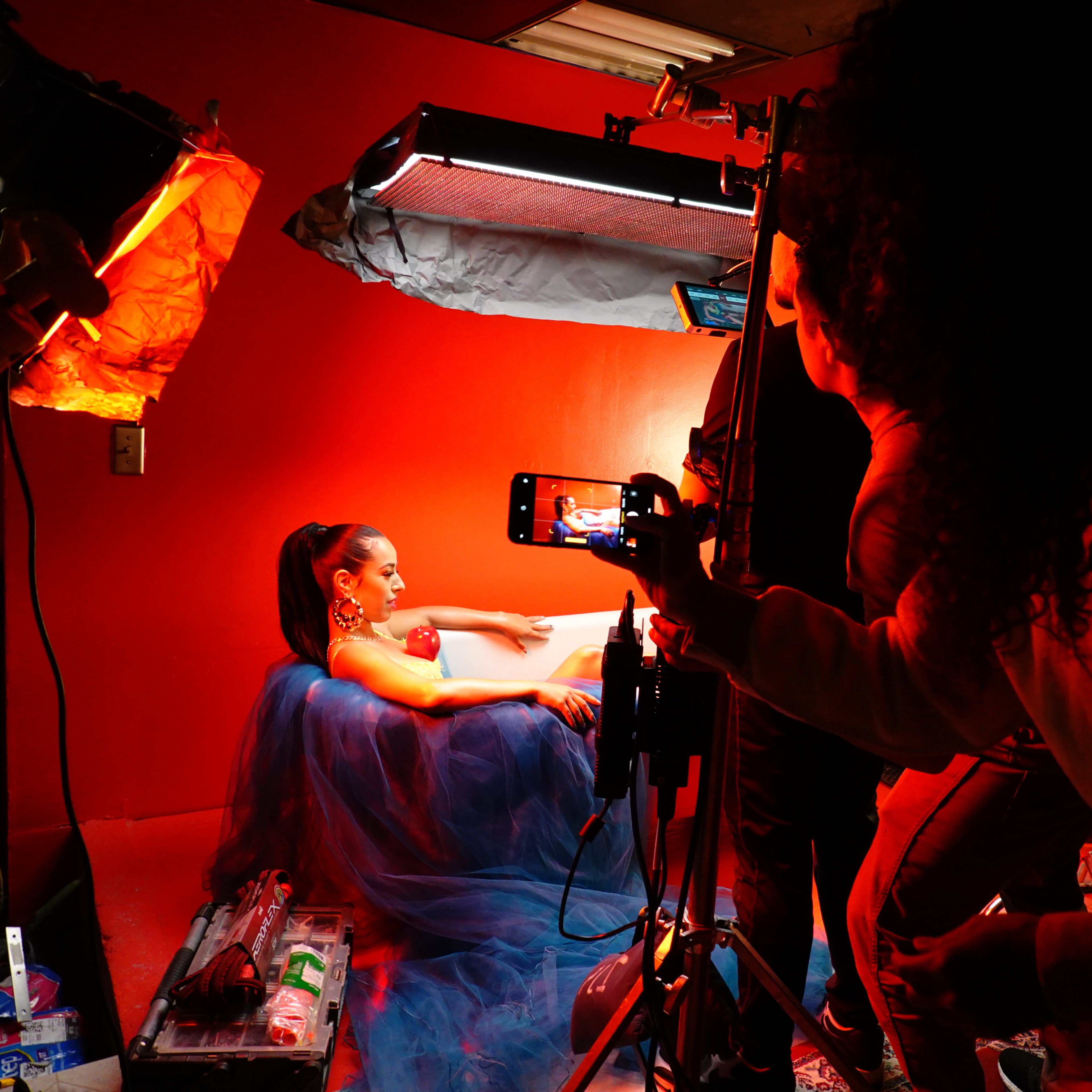 A model in a blue dress poses against a red wall while being photographed by a crew using studio lighting.