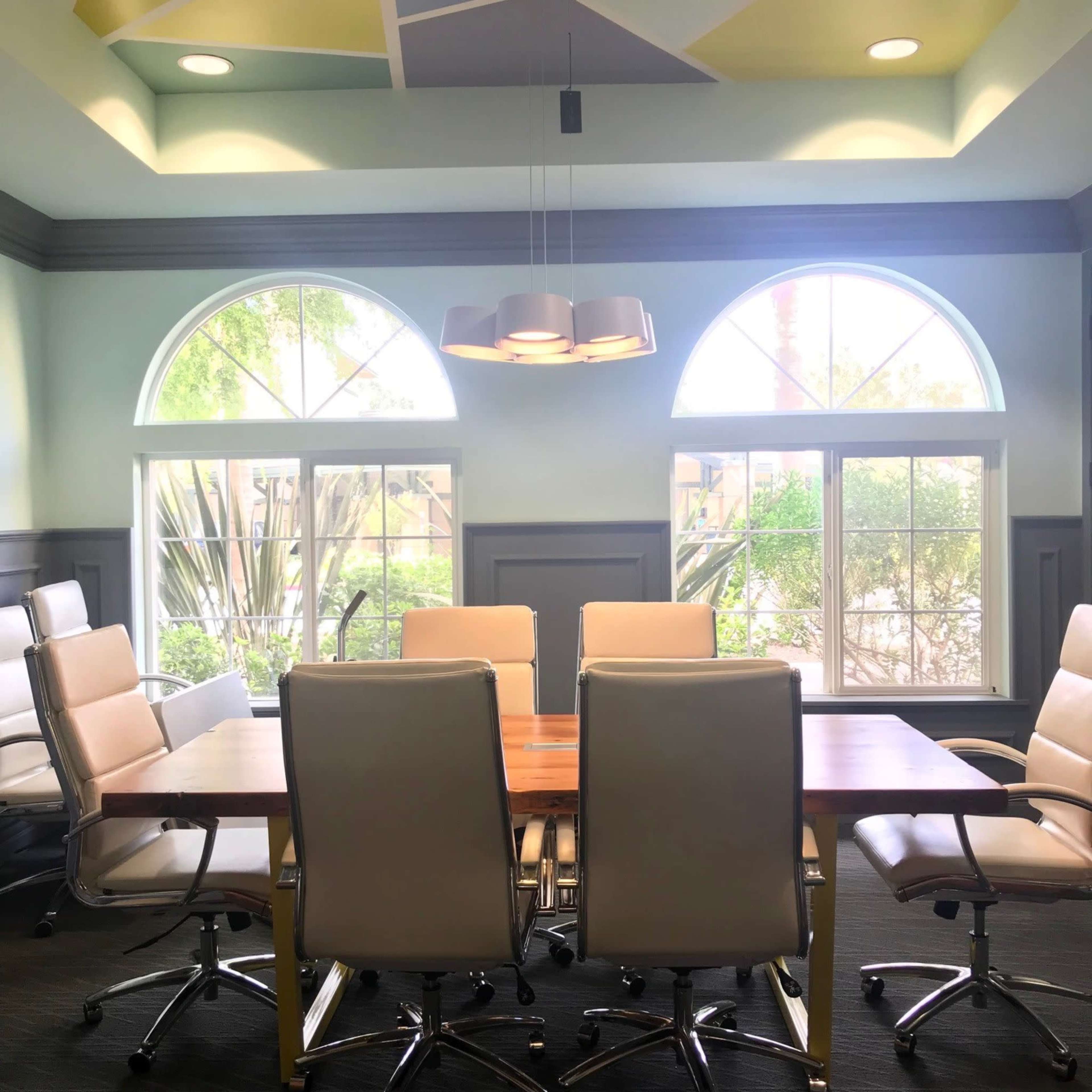 A conference room features a wooden table surrounded by rolling office chairs beneath a patterned ceiling and large windows.