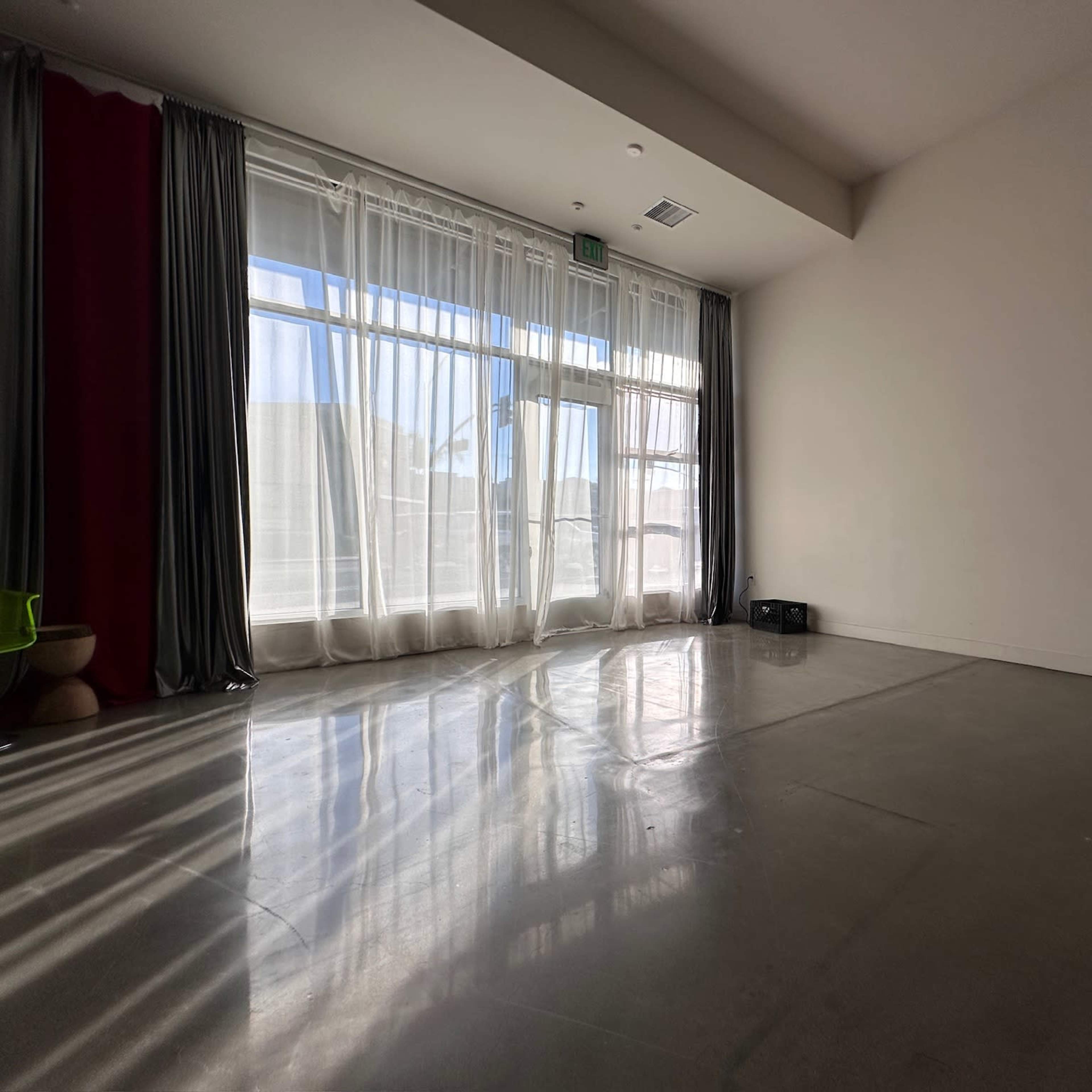 A spacious, minimally furnished room with large windows covered by sheer curtains, casting shadows on the polished concrete floor.