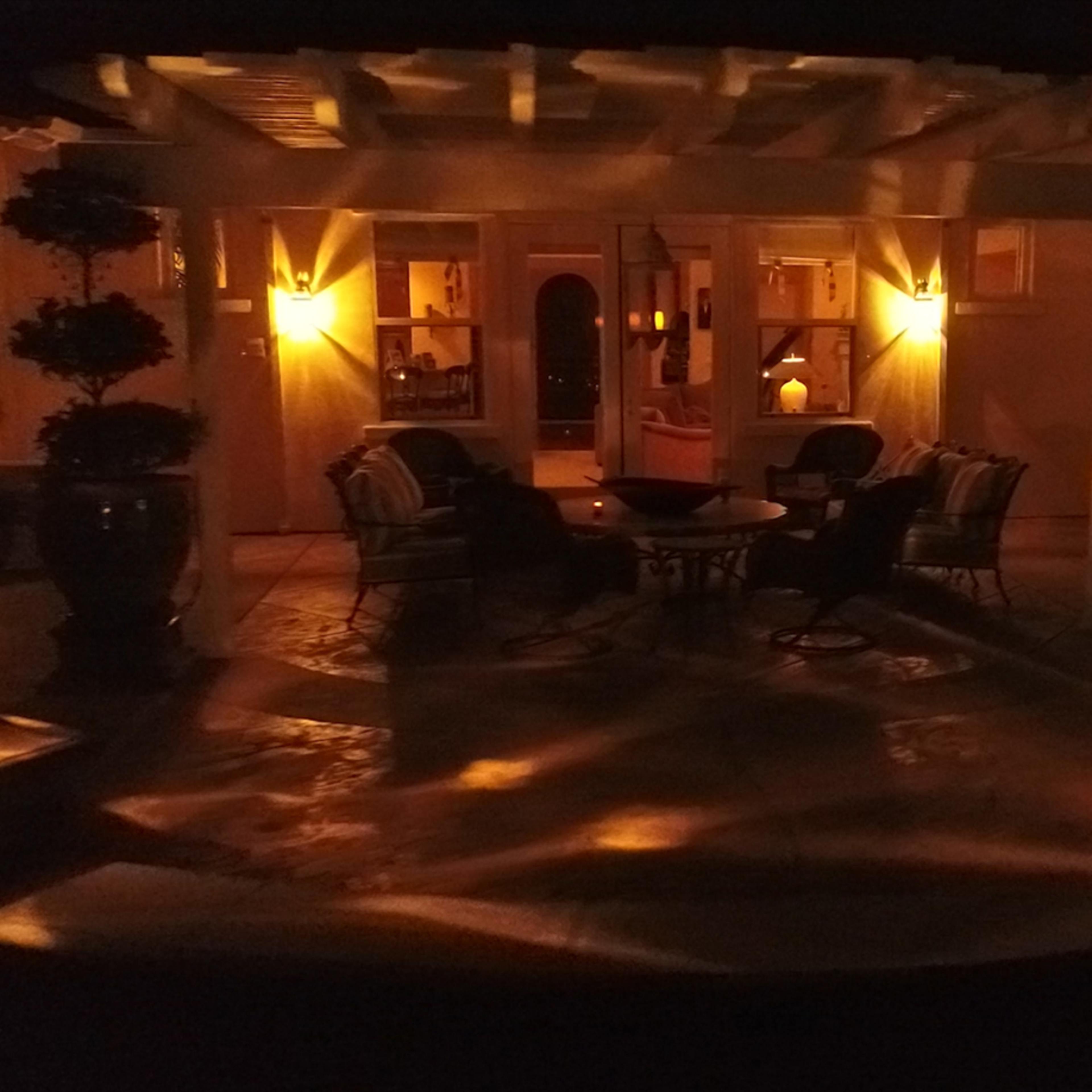 A patio area illuminated by soft lights, featuring a circular table surrounded by chairs and decorative plants.
