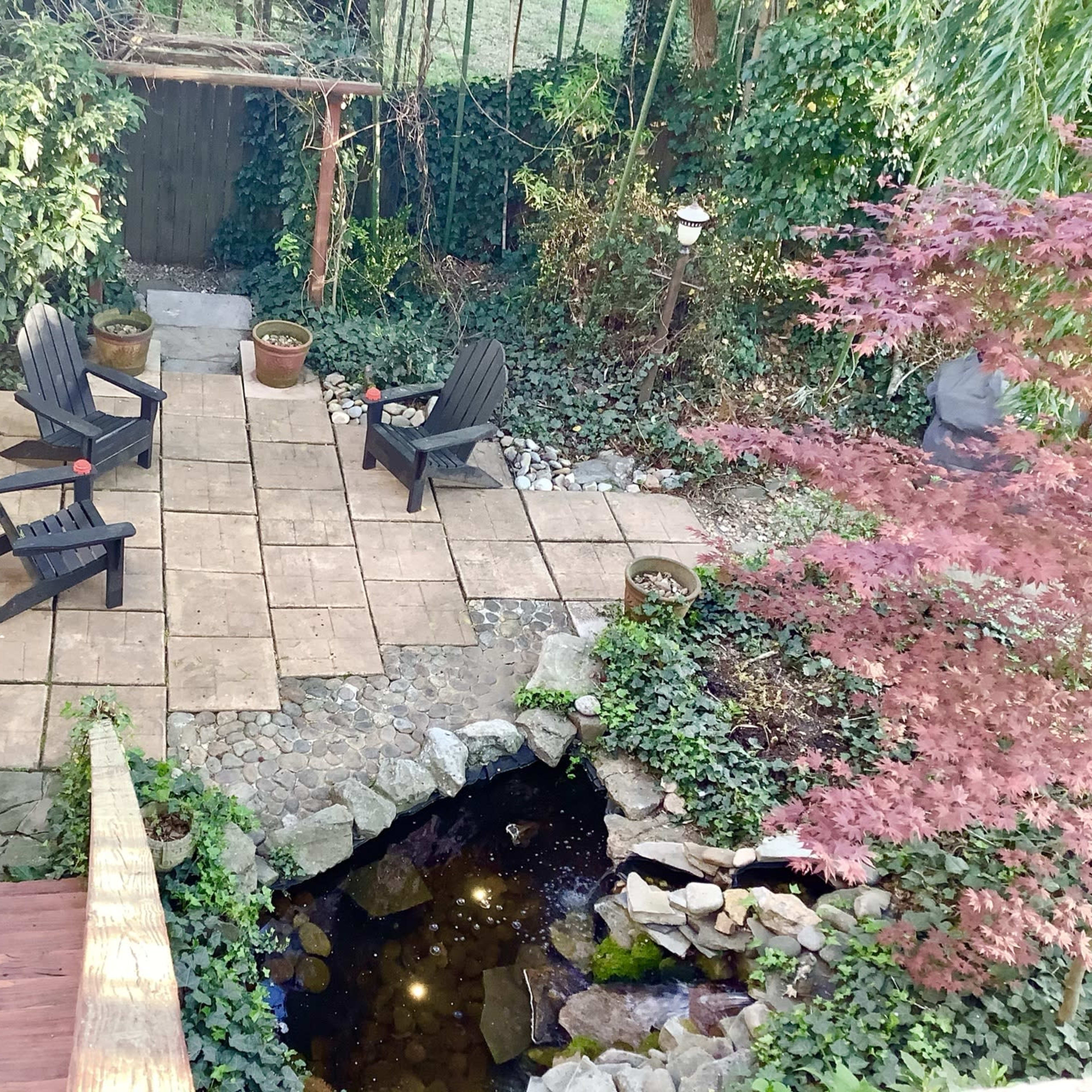 A landscaped backyard with a stone patio, two black Adirondack chairs, potted plants, and a small pond surrounded by greenery.