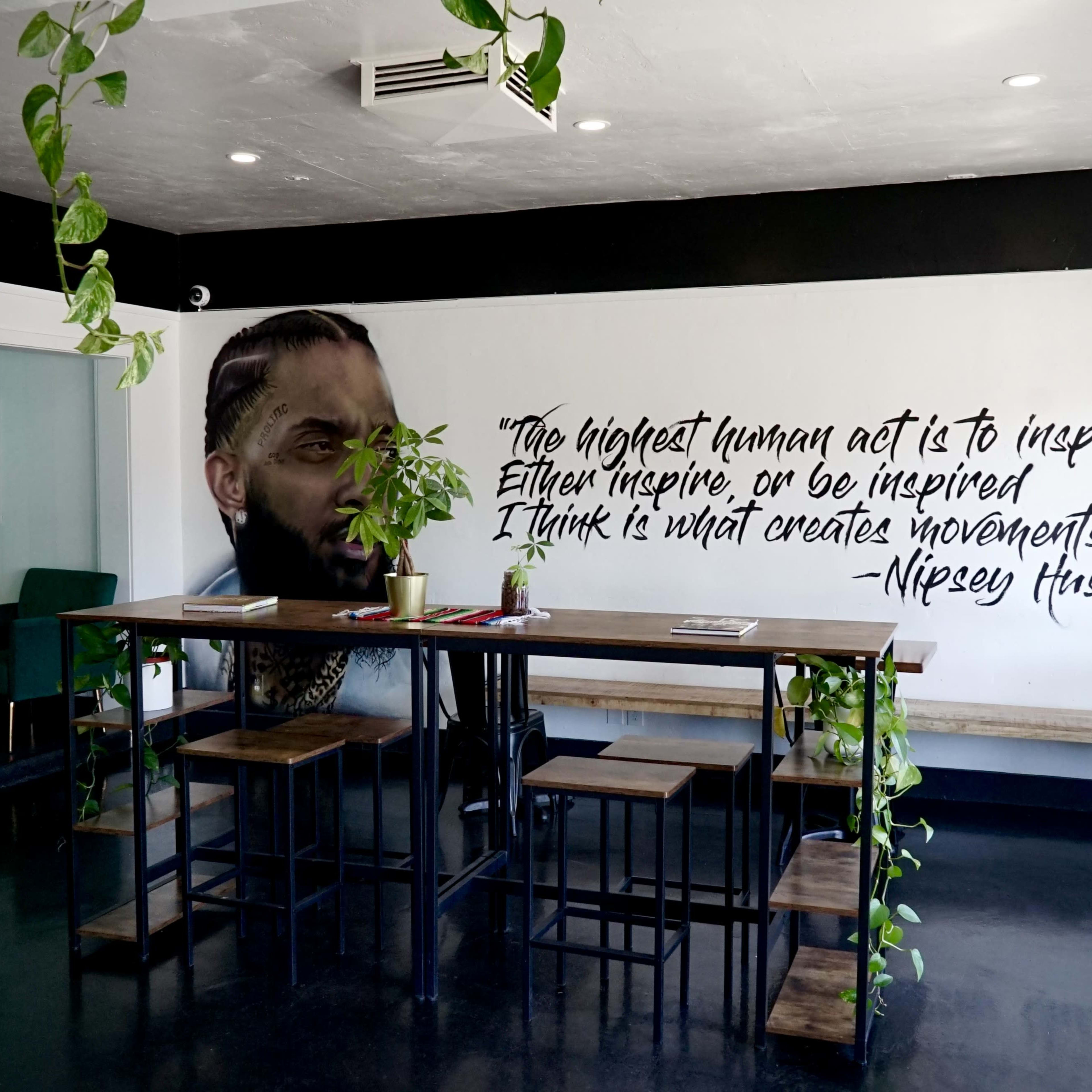 A modern café interior featuring a large mural of a man's face alongside a quote about inspiration, with a wooden table and plants in the foreground.