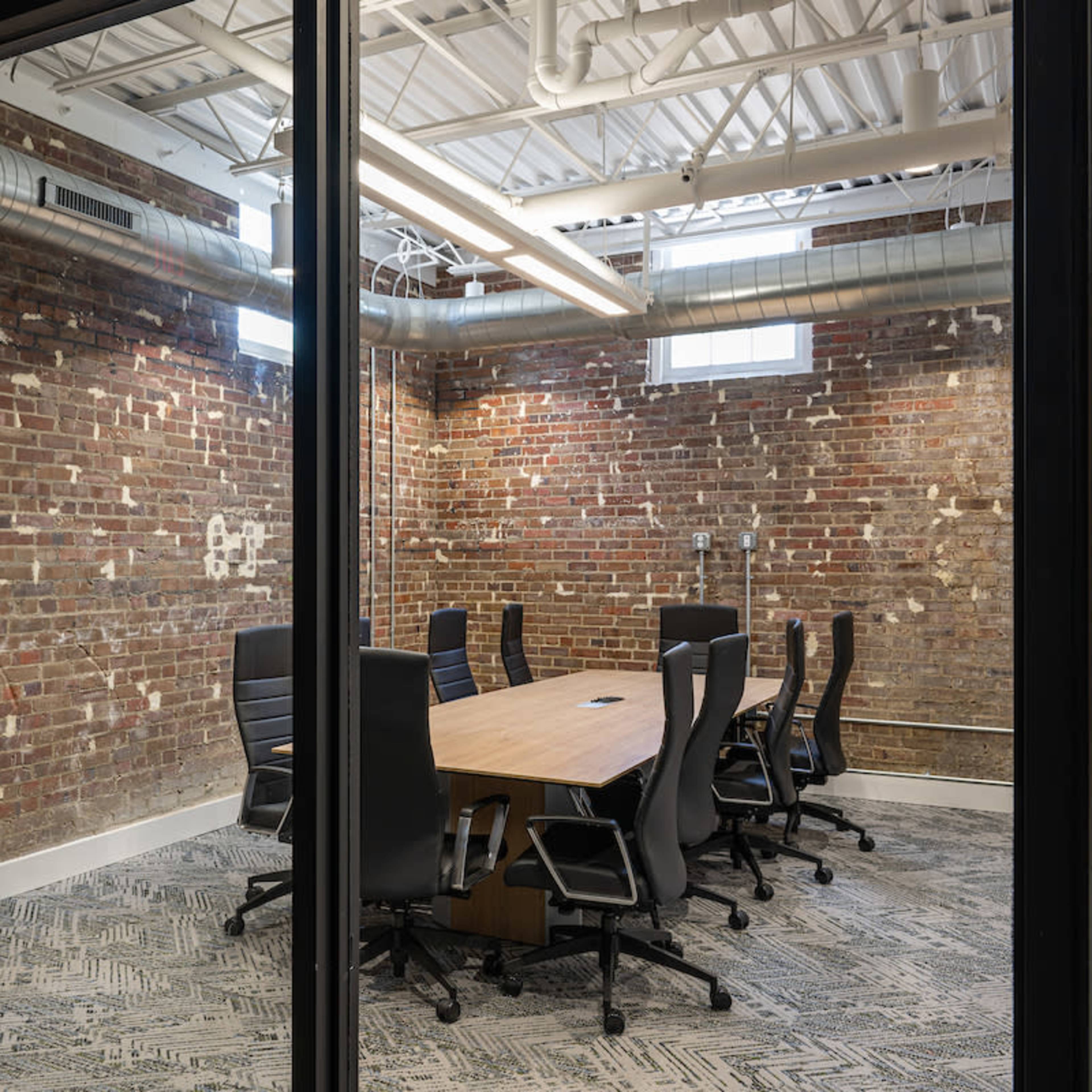 A modern conference room with a large wooden table surrounded by ergonomic chairs, set against a backdrop of exposed brick walls and a patterned carpet.