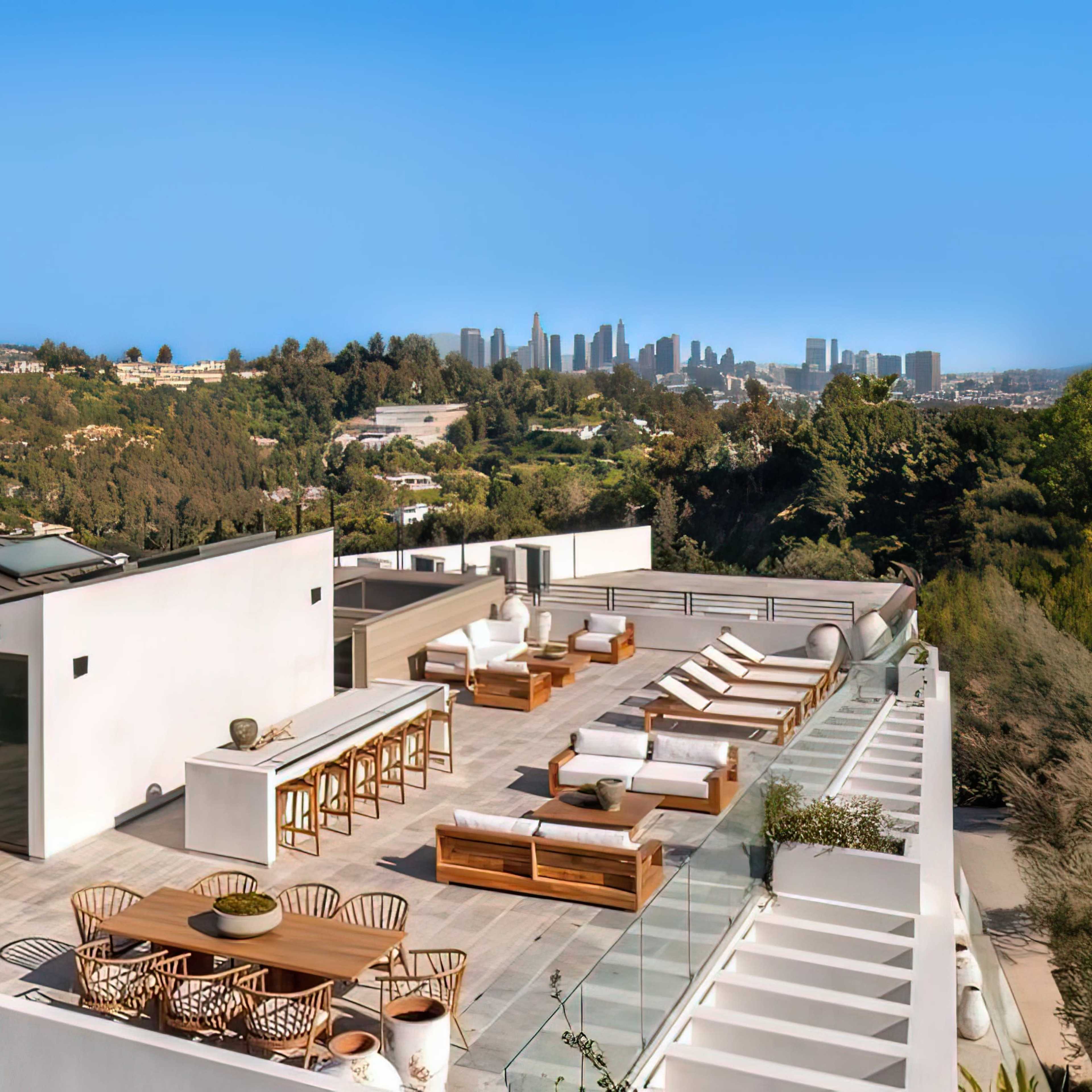 A modern rooftop terrace features dining and lounging areas with panoramic views of a city skyline and surrounding greenery.