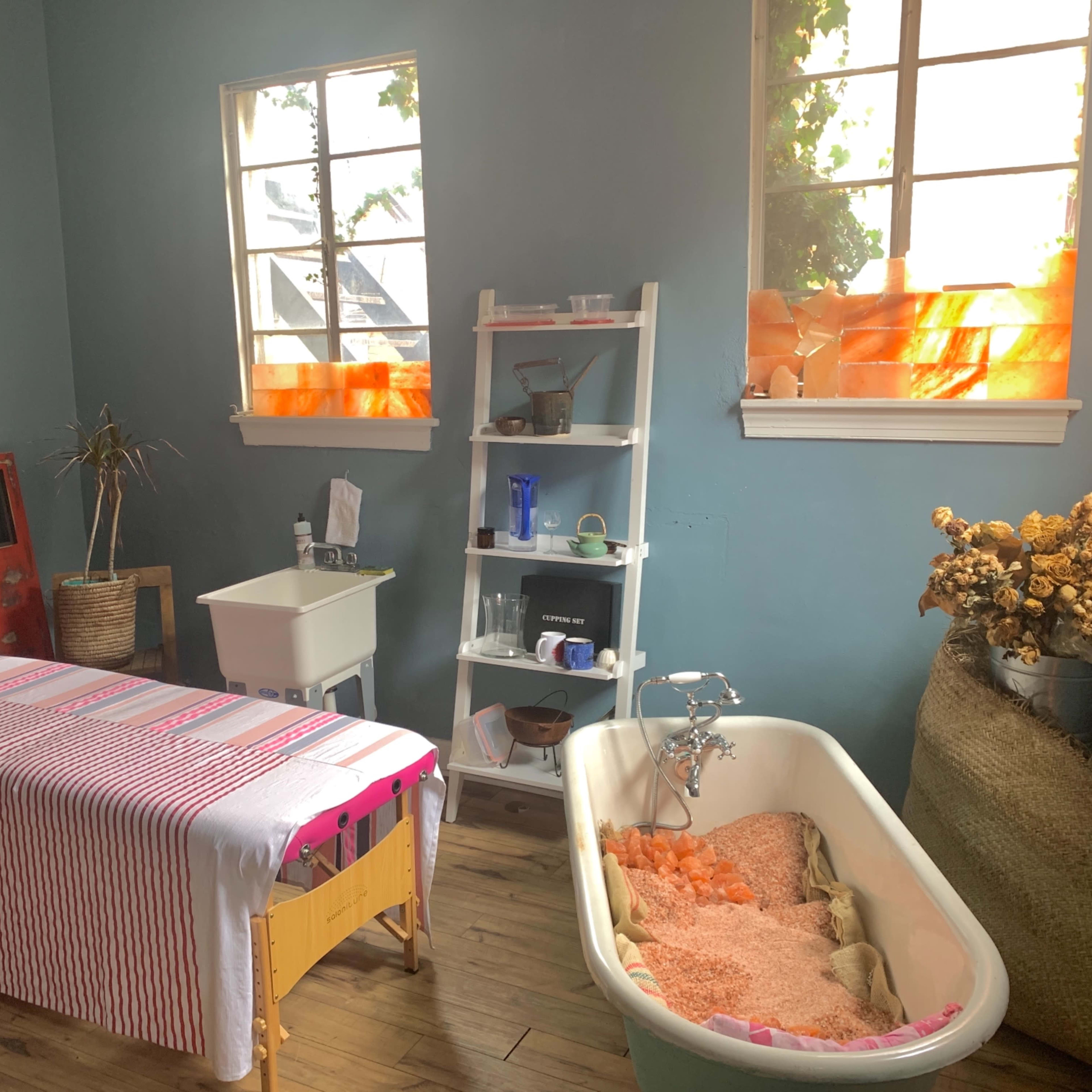 A simple room with a massage table, a vintage bathtub filled with orange bath salts, a white shelf with various items, and two windows with orange curtains.