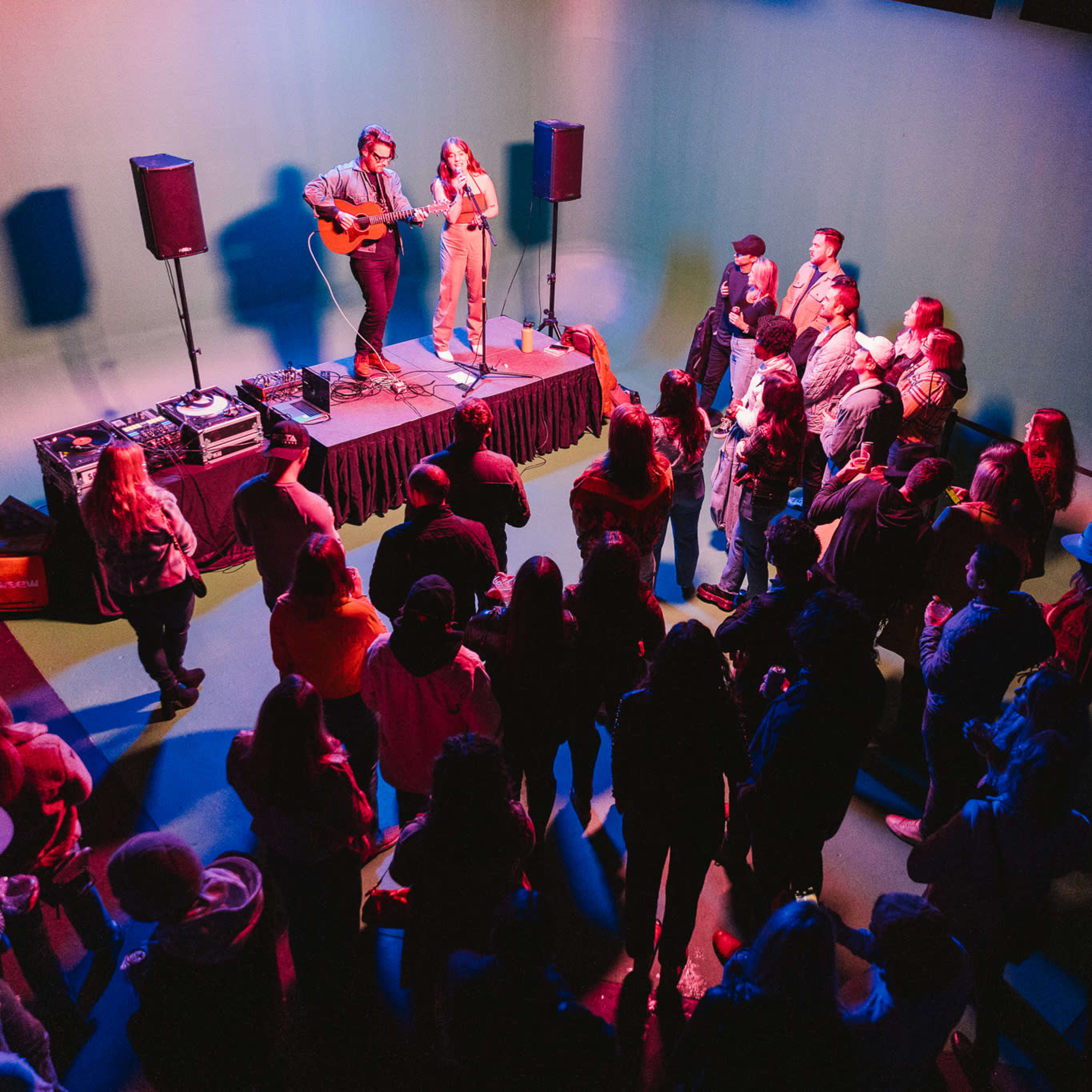 A crowd gathers around two musicians performing on a small stage with colorful lighting.
