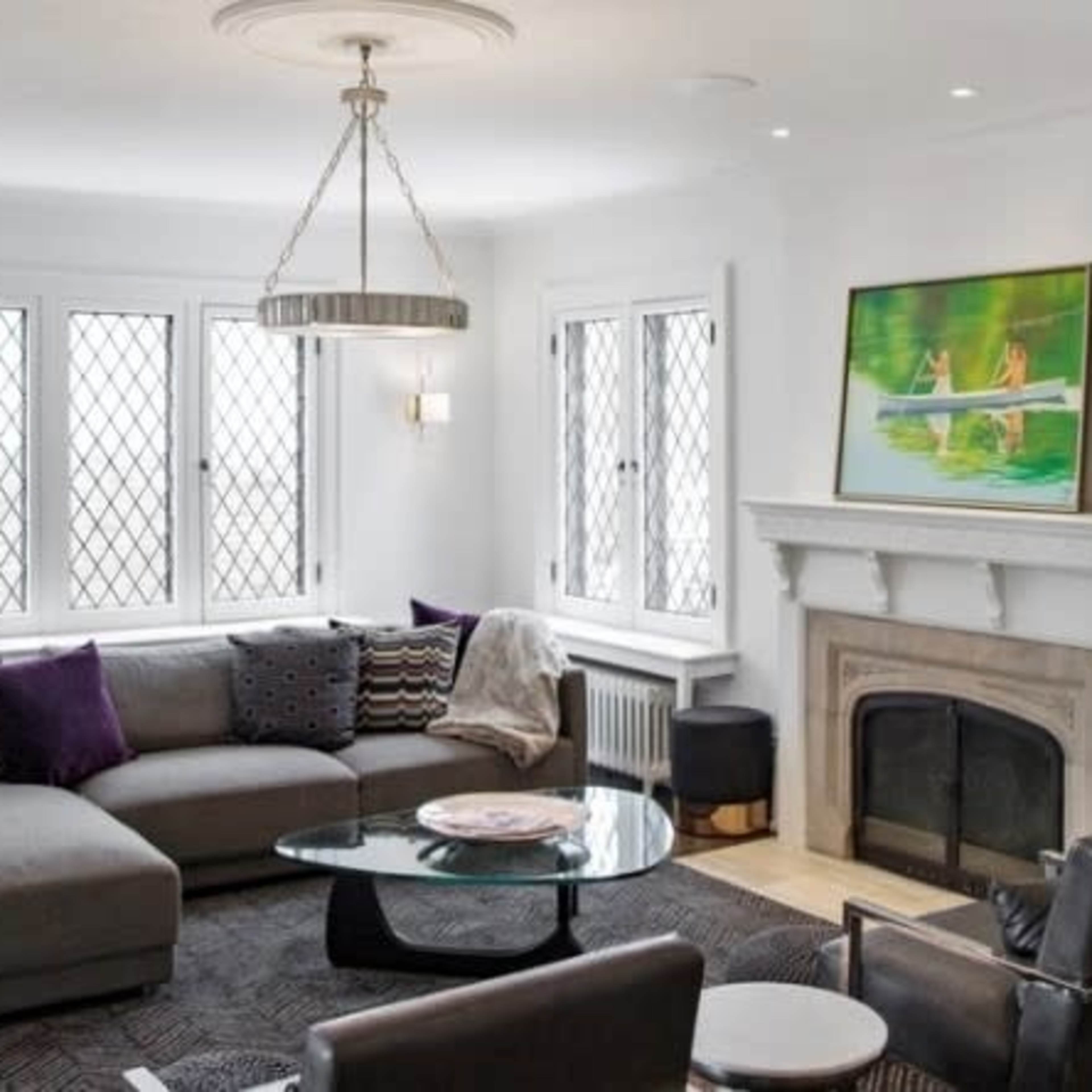 A modern living room features a gray sectional sofa, a glass coffee table, and a fireplace beneath a decorative painting, all set against a backdrop of large diamond-patterned windows.