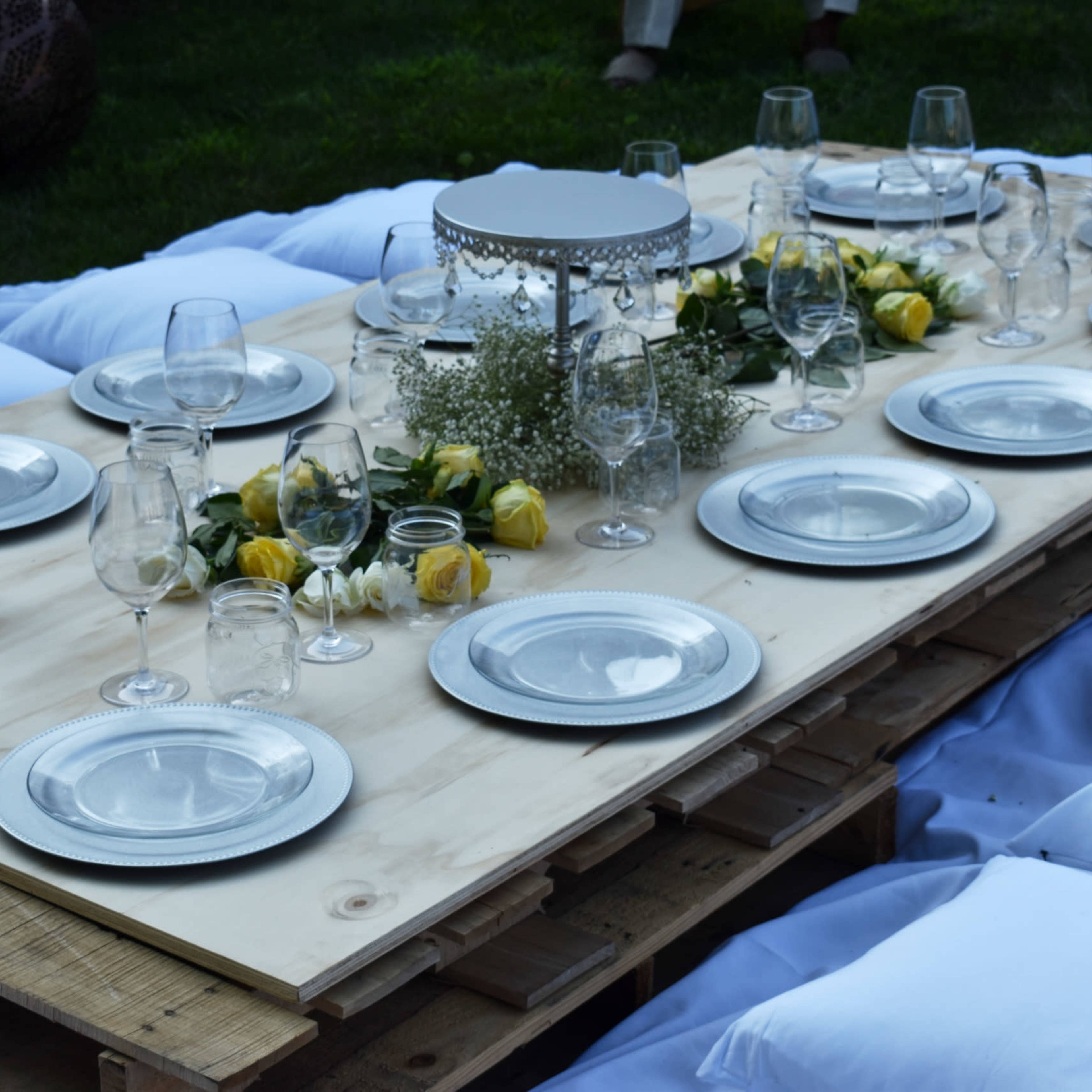 A wooden table is set with plates, glasses, and floral decorations on a grassy area, surrounded by large pillows for seating.