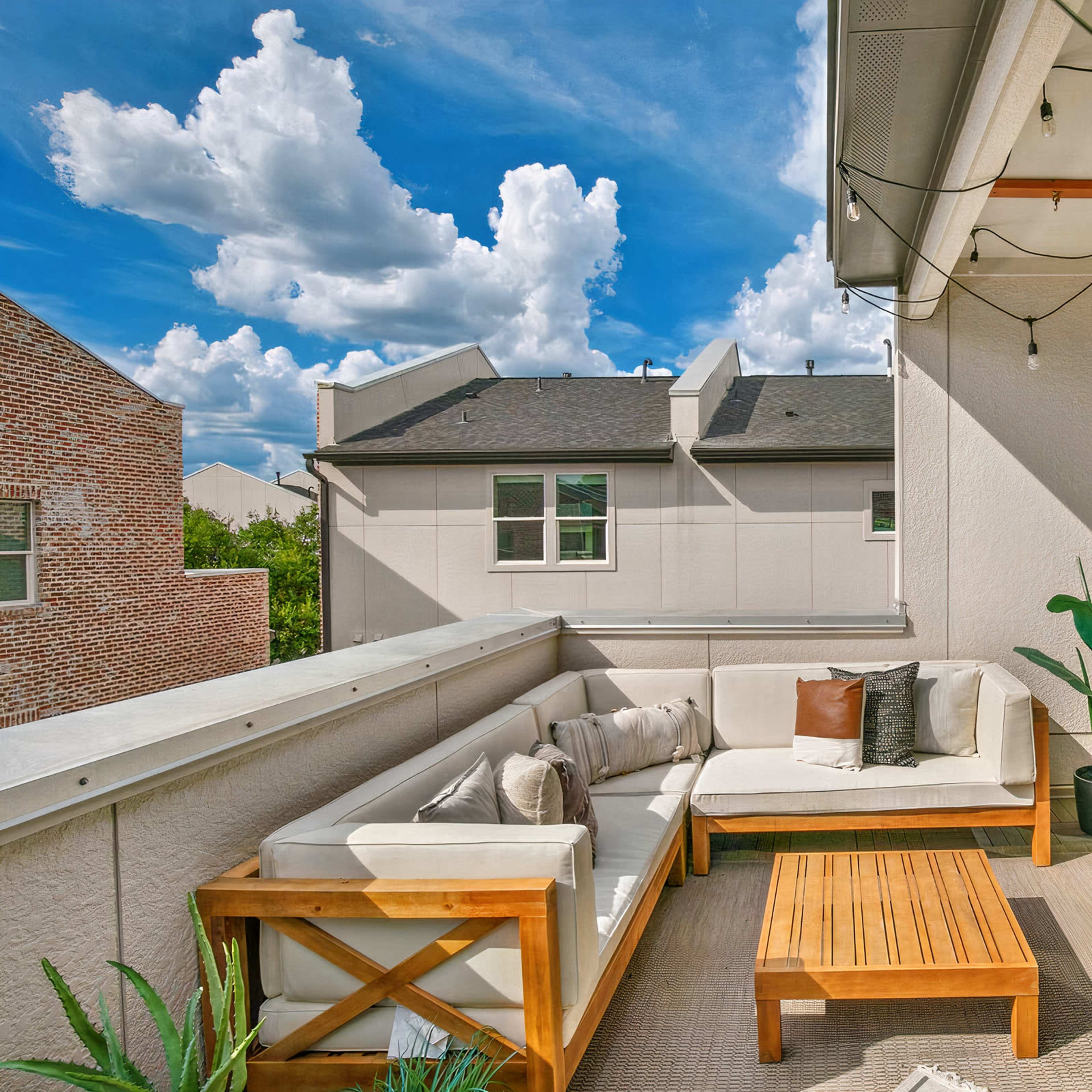 A balcony with a wooden seating area, a hanging hammock, and potted plants, set against a backdrop of blue sky and clouds.