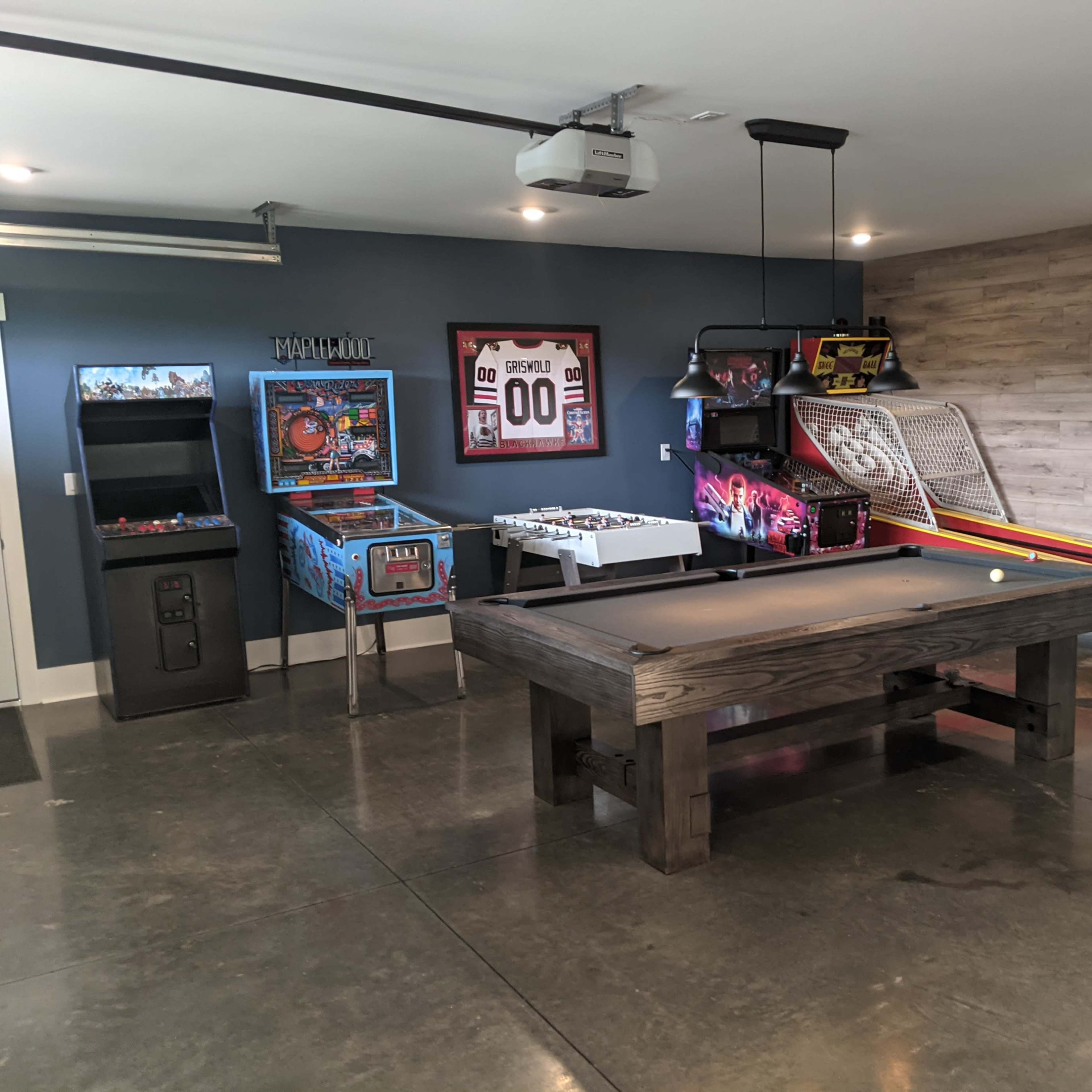 A game room features a billiards table, arcade machines, and a skee ball setup against a gray wall.