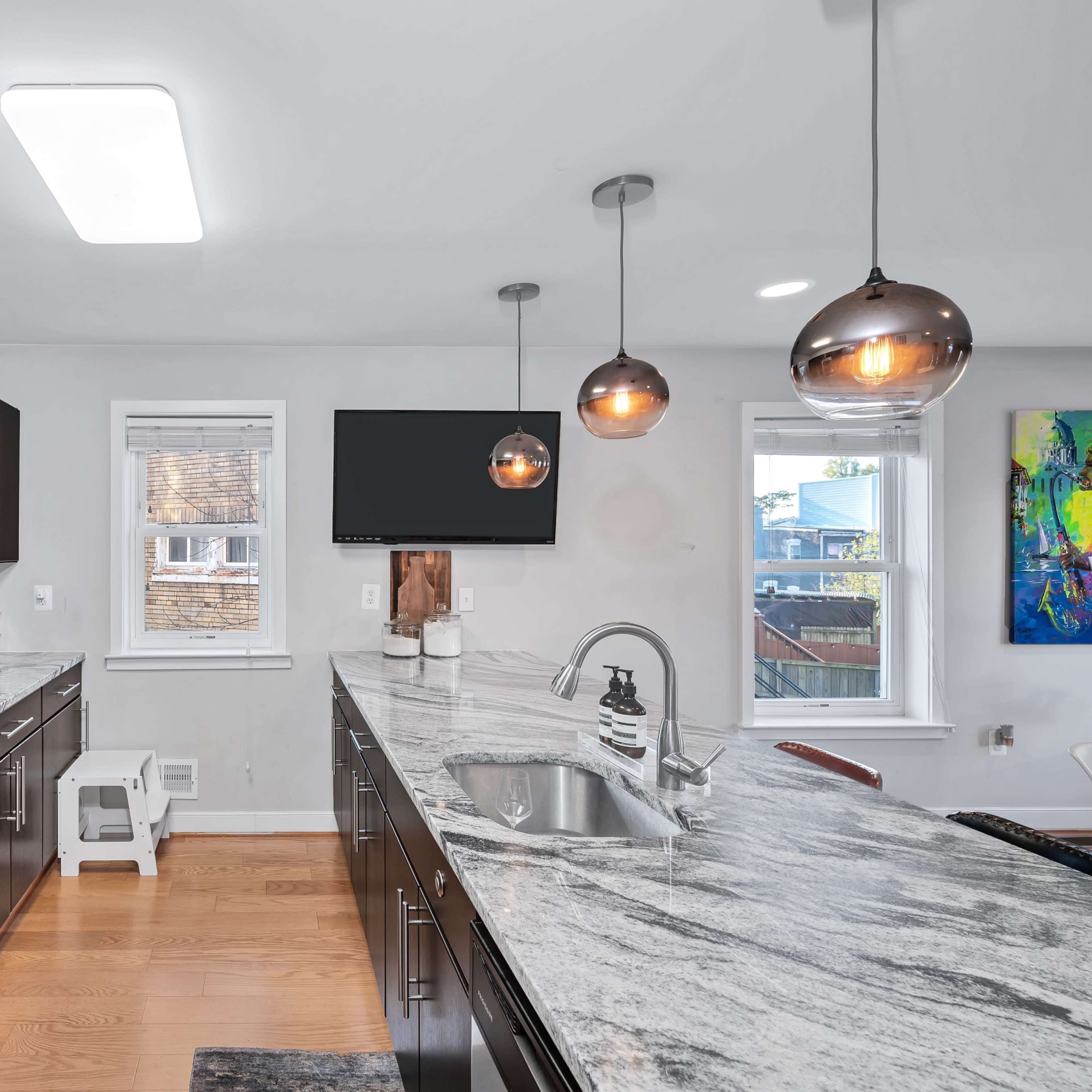 A modern kitchen features dark cabinetry, stainless steel appliances, and a large marble countertop with pendant lighting above.