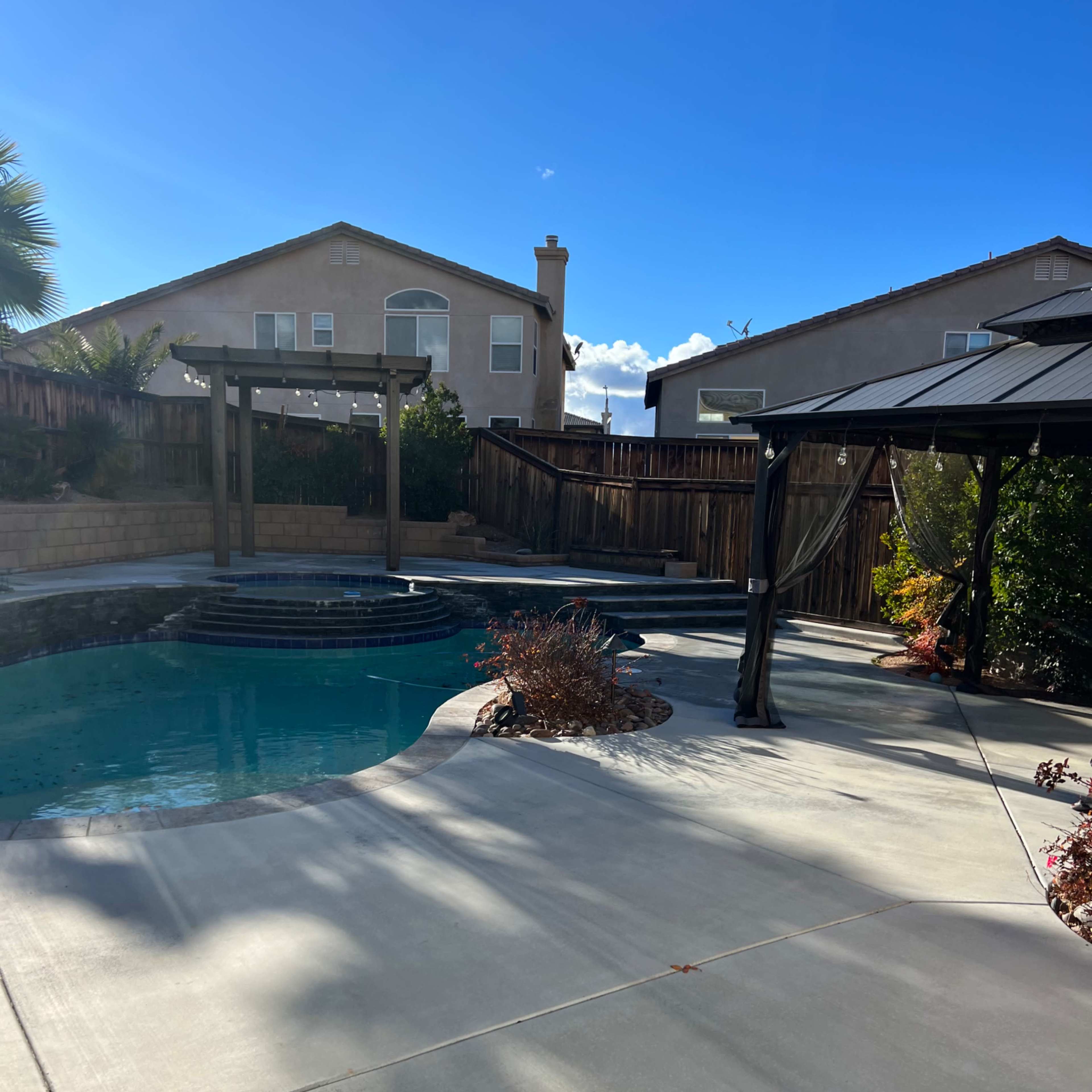 A backyard features a pool with a stone edge, surrounded by landscaping and a shaded gazebo under a clear blue sky.