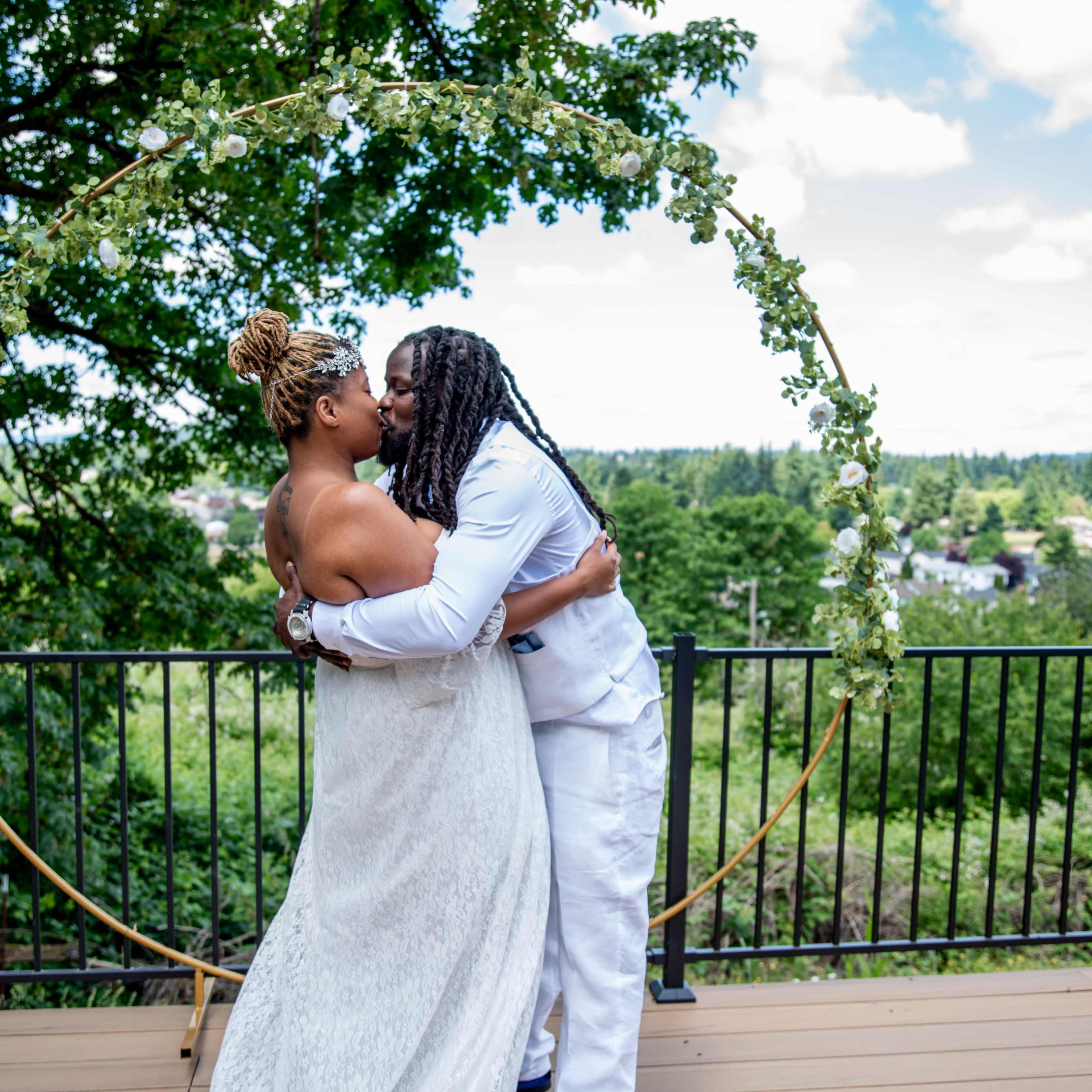 A couple kisses under a floral arch with a scenic background of trees and clouds.