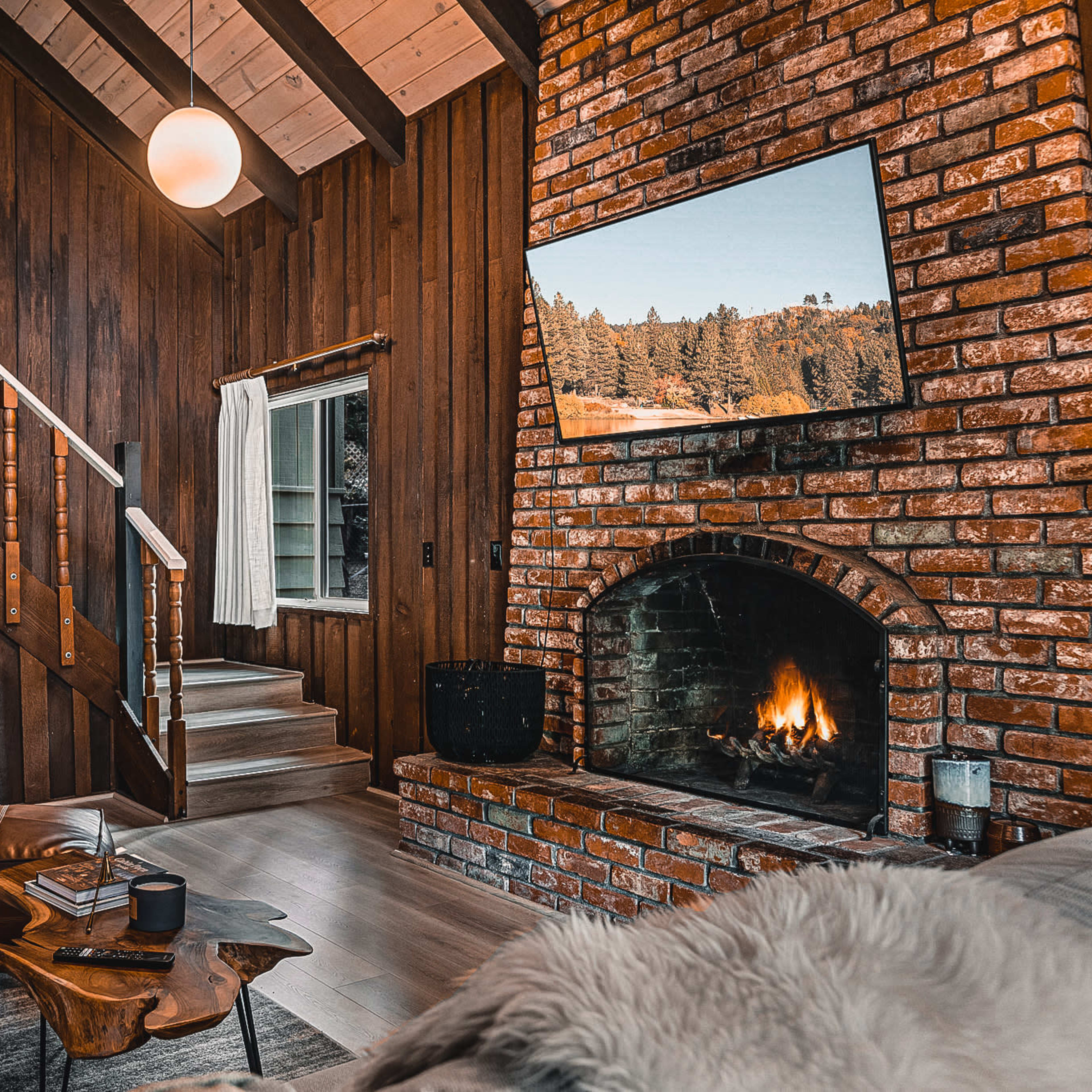 The interior of a rustic living room features a brick fireplace, a television mounted on the wall, wooden paneling, and a staircase leading to an upper level.