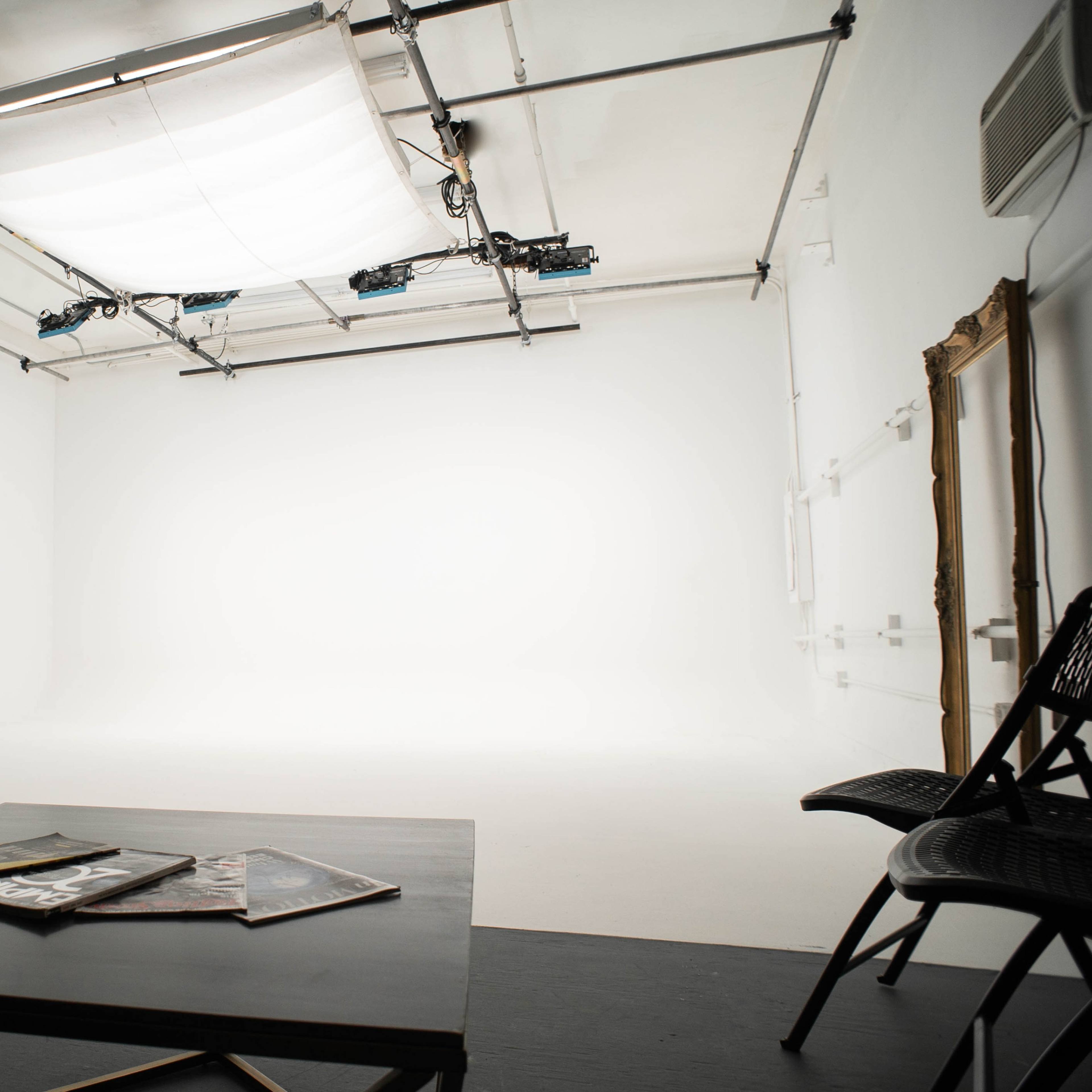 A minimalist studio space with a large white backdrop, a low table with magazines, and a chair positioned against the wall.