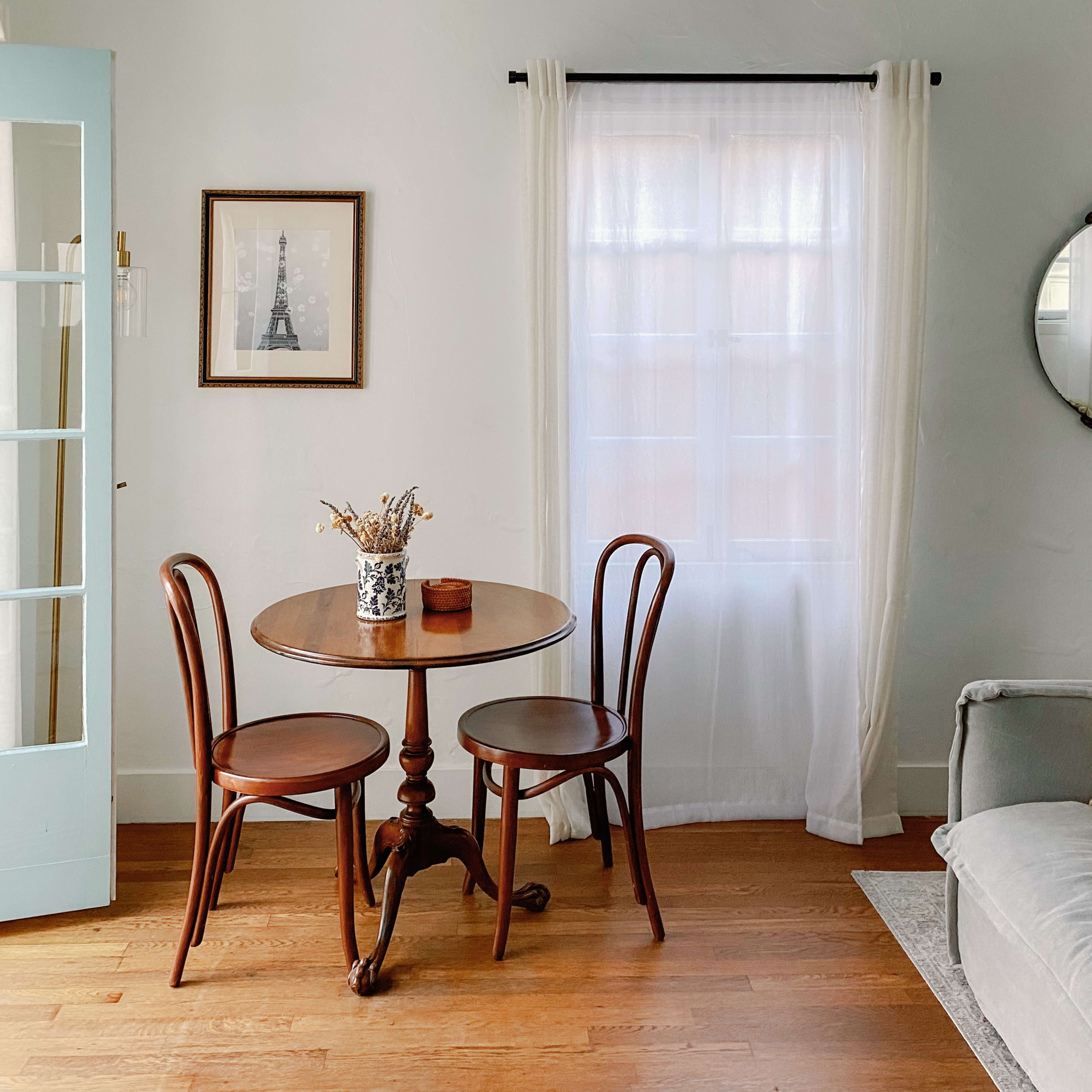 A small dining table with two chairs is positioned next to a window in a well-lit room featuring a gray sofa and a round mirror on the wall.