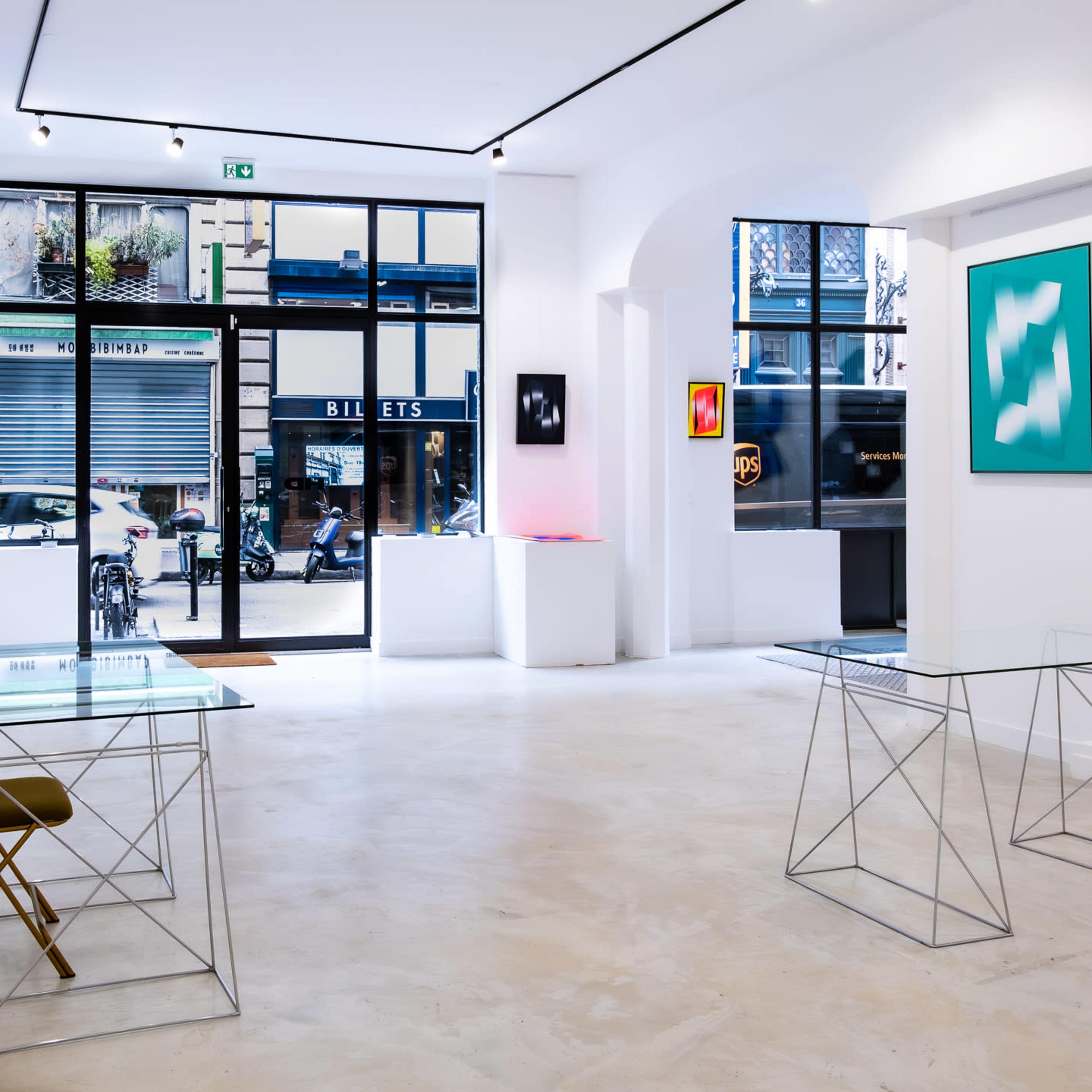 A contemporary art gallery featuring two glass-topped tables with metal bases, a couple of framed artworks on the walls, and large windows allowing natural light to enter.