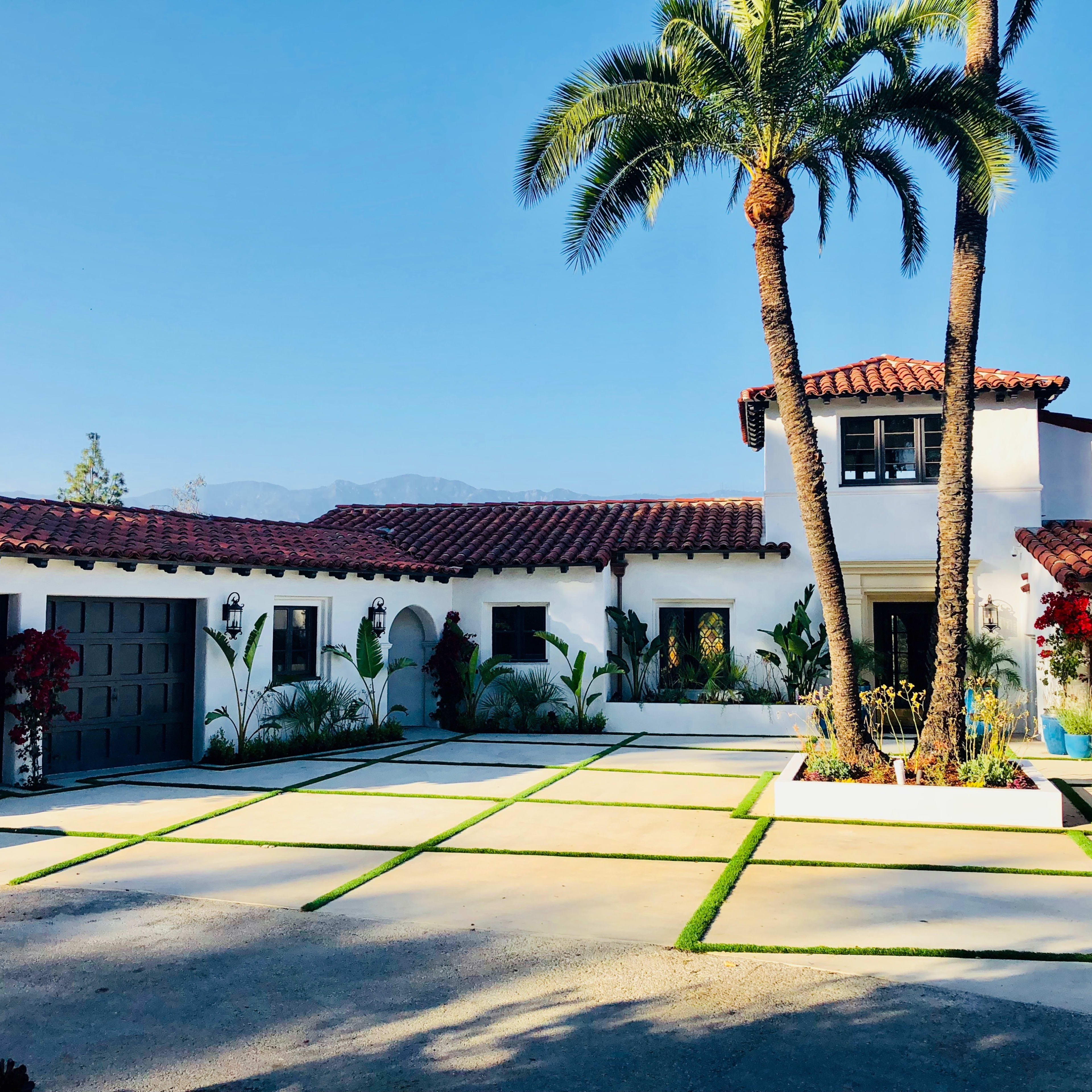 A Mediterranean-style house with a tile roof is set in a landscaped courtyard featuring palm trees and decorative plants.