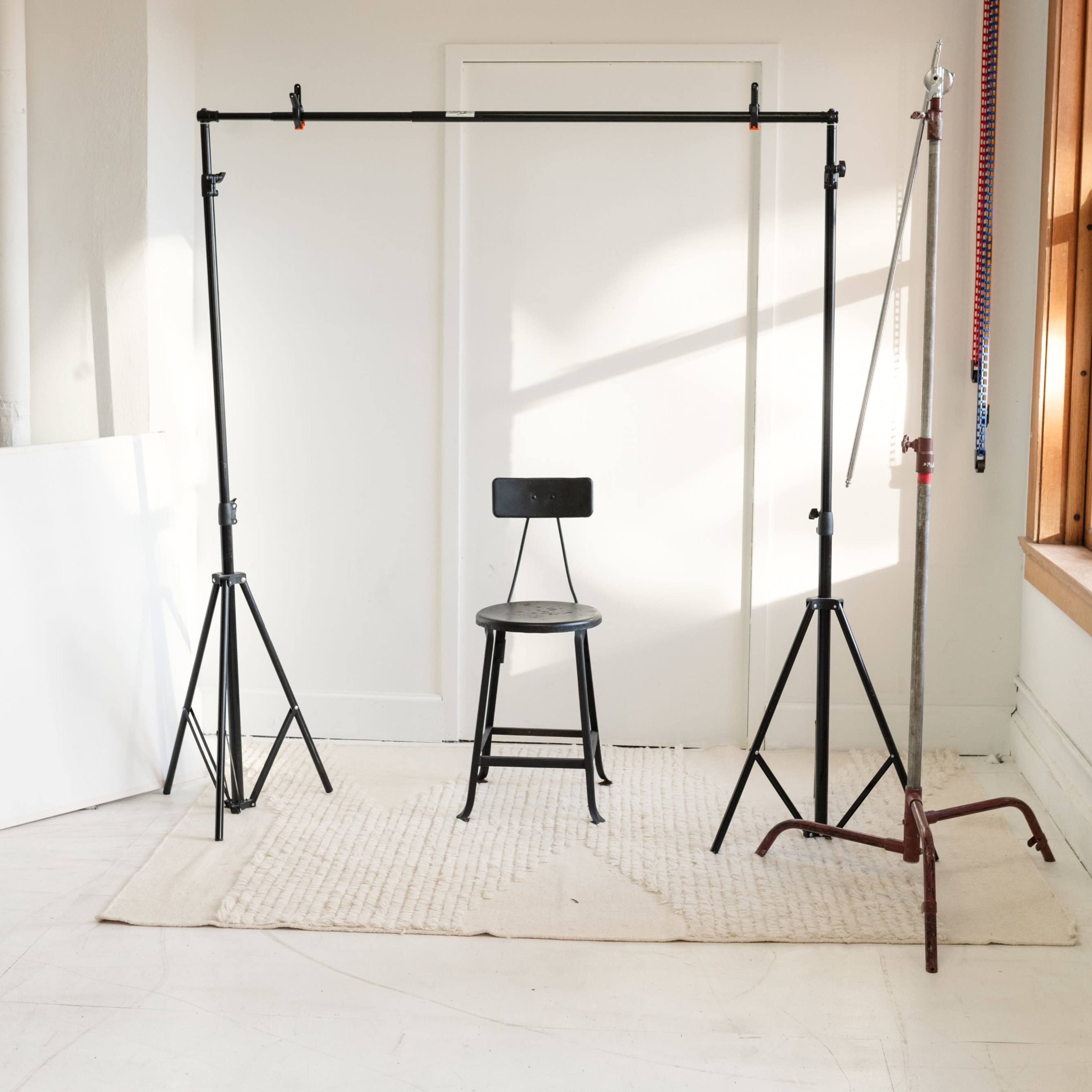 A black stool is positioned in front of a backdrop setup made of pipe stands in a brightly lit room with wooden windows.