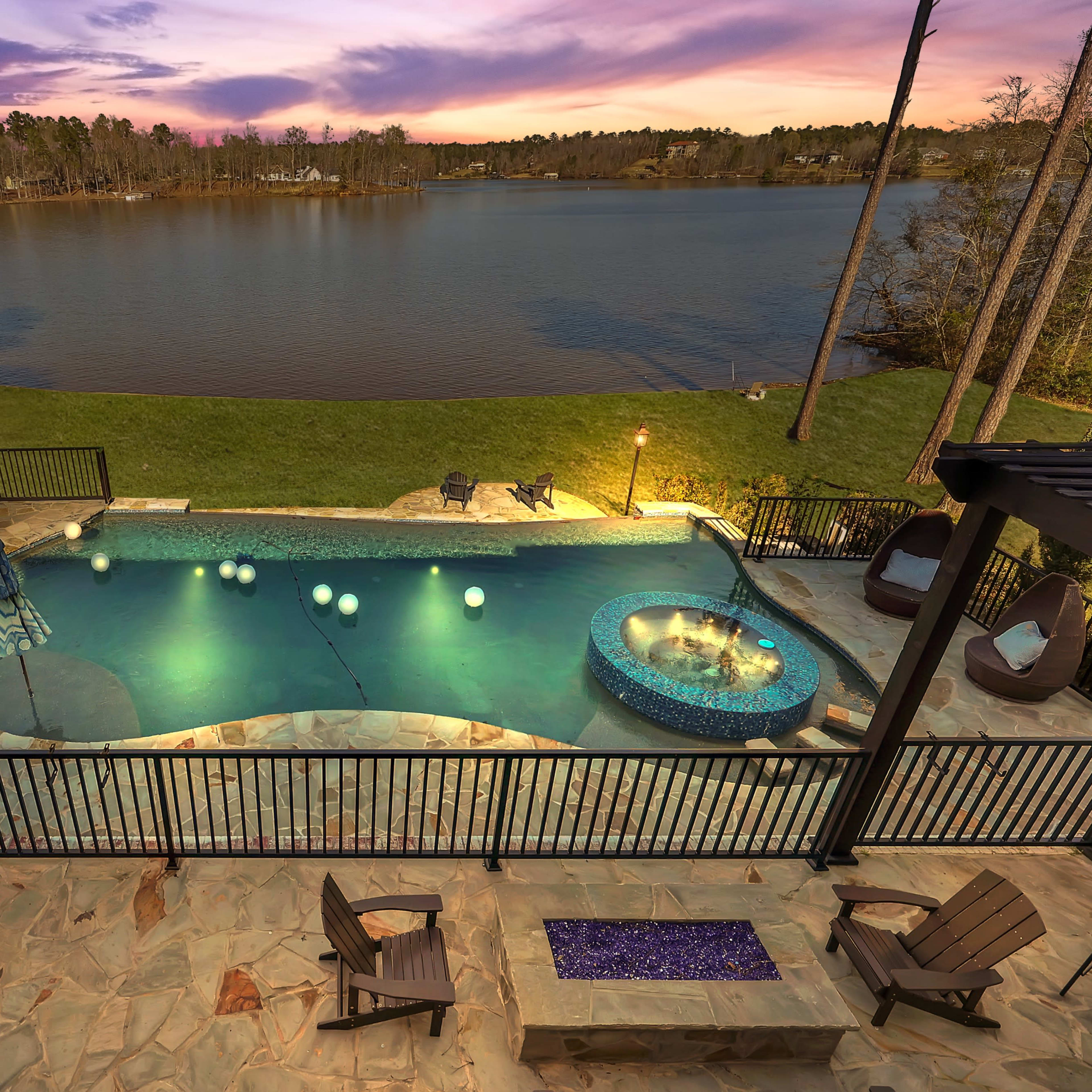 A landscaped outdoor area with a swimming pool, spa, and a view of a lake at sunset.