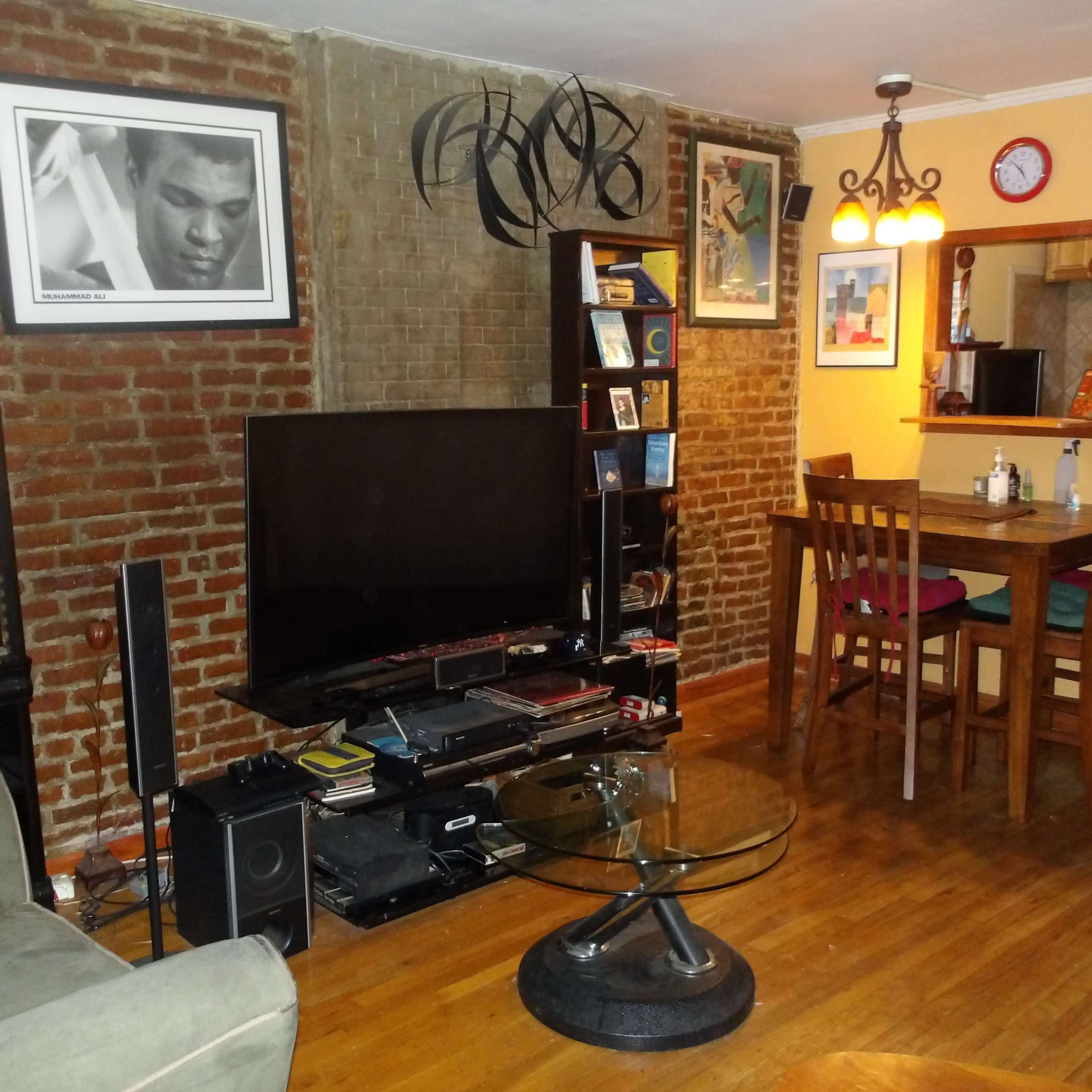 A cozy living room with exposed brick walls, a large TV on a stand, a bookshelf filled with books, a circular glass coffee table, and a dining area with a wooden table and chairs.